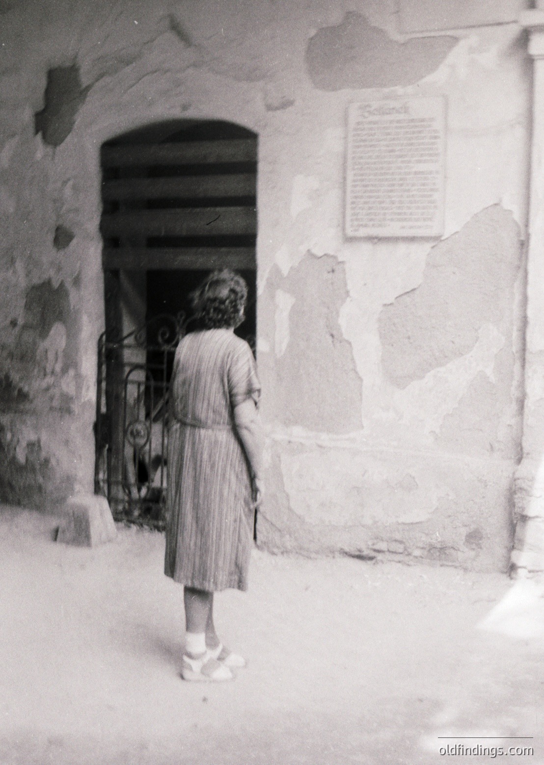 Mid-20th century girl in pleated dress stands at weathered stone doorway with wrought-iron gate. Plaque on wall suggests historical or cultural significance. Rustic, likely European architecture.