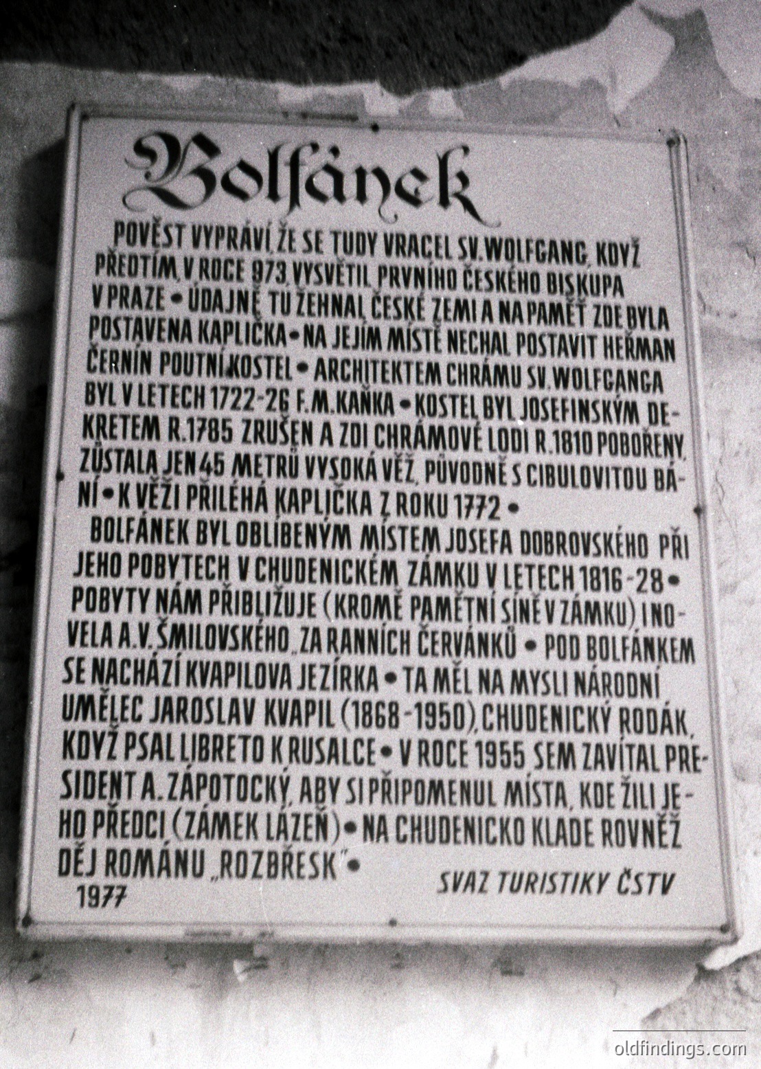 A black-and-white commemorative plaque mounted on a stone wall, detailing historical events in **Bolfánek**, Czech Republic. The text references St. Wolfgang’s healing in 973, the 1722–26 construction of a church by F.M. Kanka, and the 1955 visit of President A. Zapotocký. Engraved in Czech, with a 1977 date from the Czech Tourist Association.