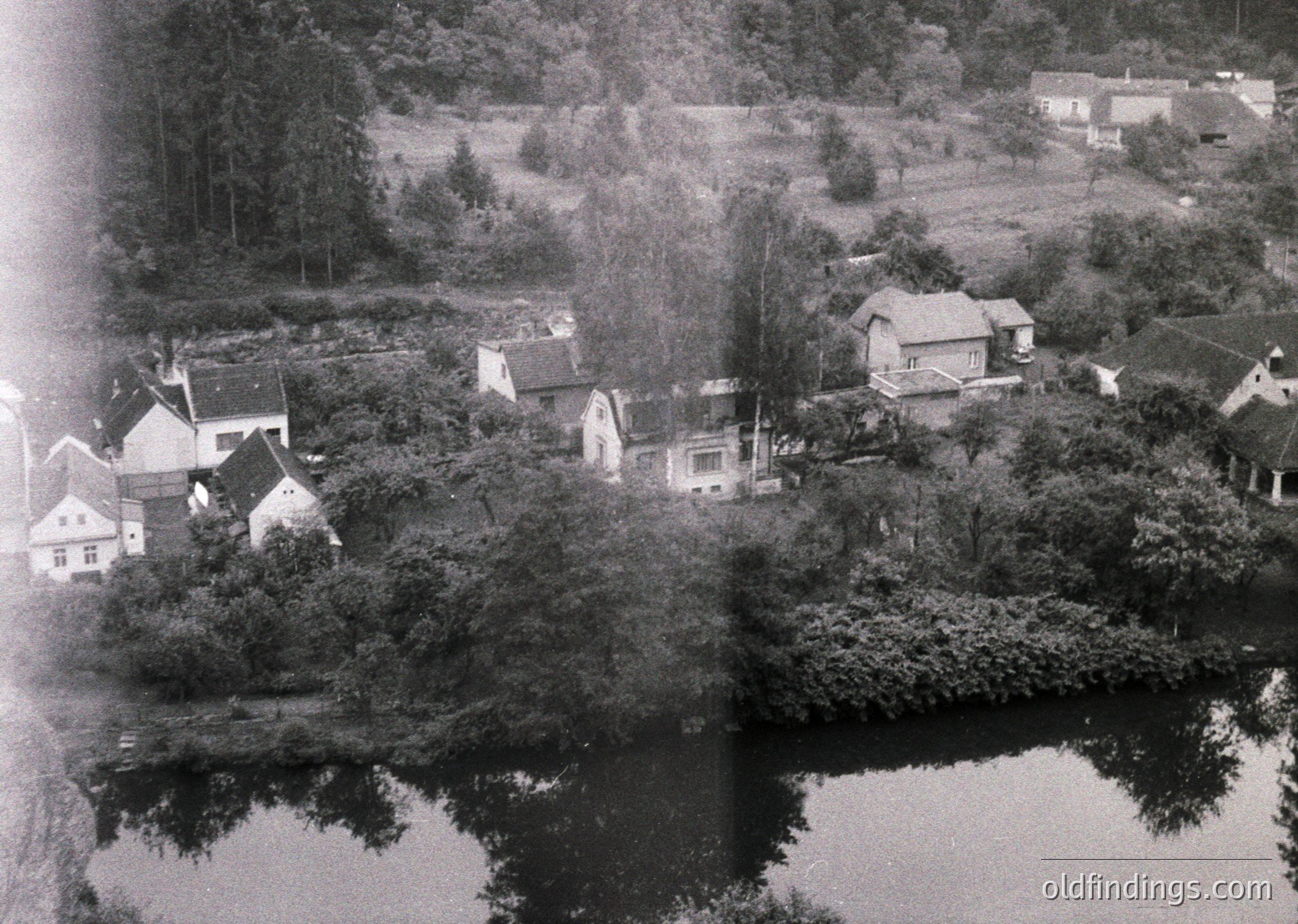 Aerial black-and-white photo of a lakeside village, likely mid-20th century. Clustered houses with pitched roofs and dense greenery surround a calm body of water, reflecting trees and structures. Rural setting with mixed residential and agricultural land.