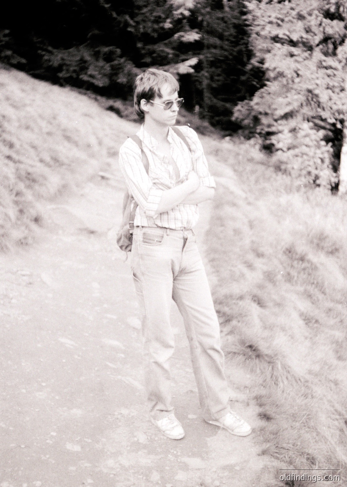 Black-and-white portrait of a person in outdoor rugged terrain, likely a coastal or desert cliffside. Mid-century casual attire: belted blouse, wide-leg trousers, and round glasses. Crossed arms pose suggests confidence.