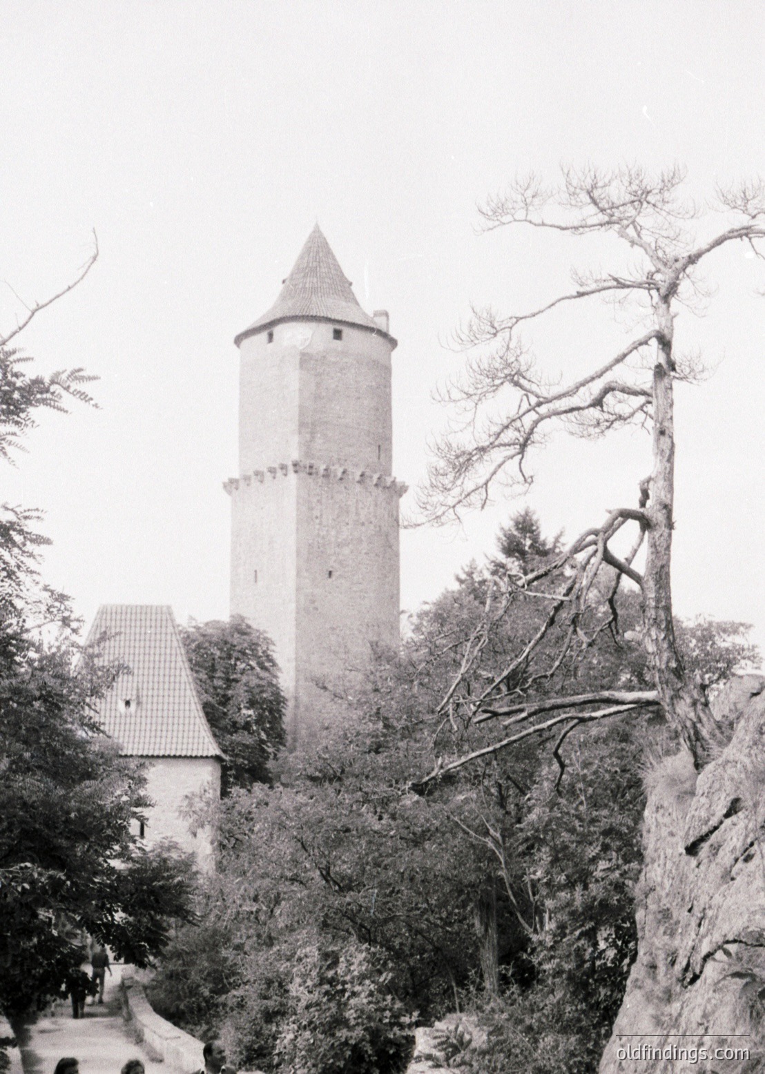 Historic stone watchtower with conical roof, surrounded by dense foliage and rocky terrain. Likely Eastern European medieval architecture, featuring crenellated upper sections.