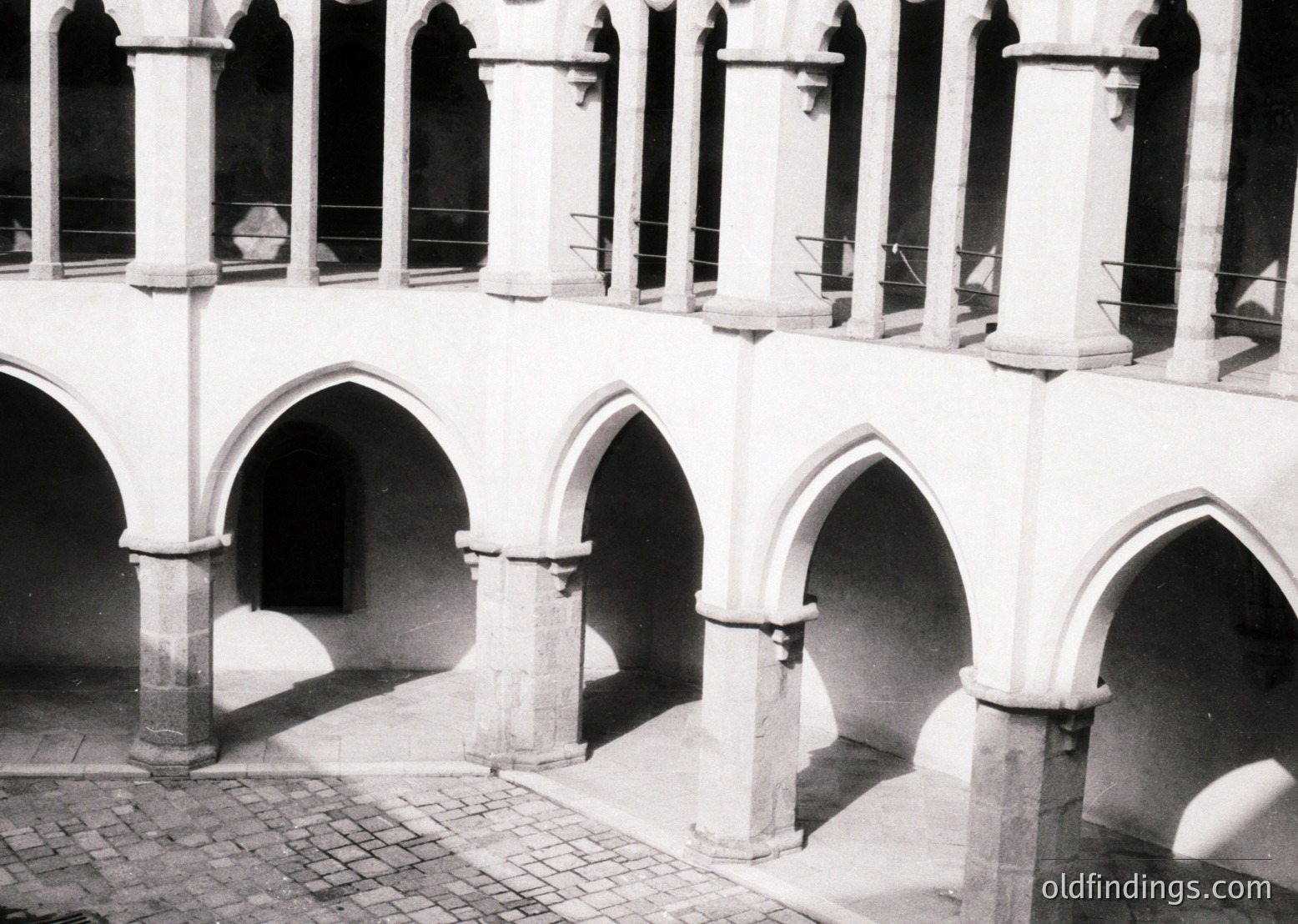 Gothic-style cloister arcade with pointed arches, stone columns, and metal railings. Likely European monastic or university architecture, mid-19th to early 20th century. Cobblestone courtyard floor with minimal wear.