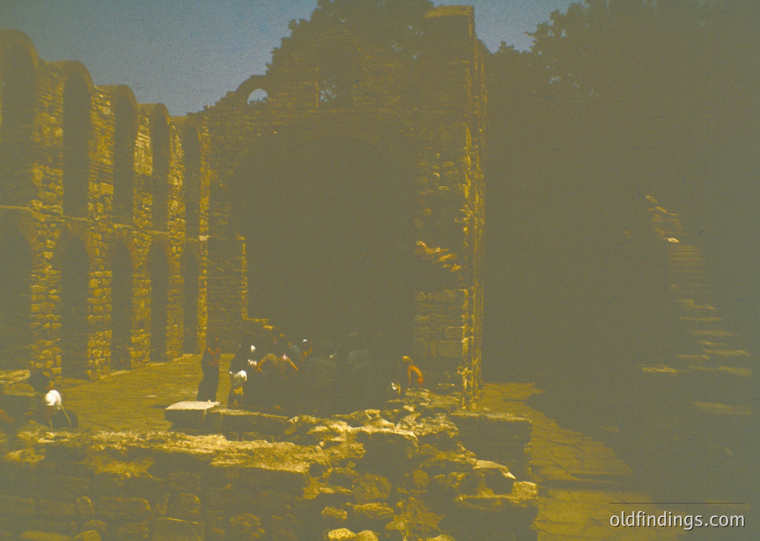 Vintage sepia-toned photograph of ancient stone ruins with weathered columns and arched doorways, likely from a Southeast Asian temple complex. Two figures in traditional attire stand near a central stone pedestal, surrounded by uneven stone flooring.