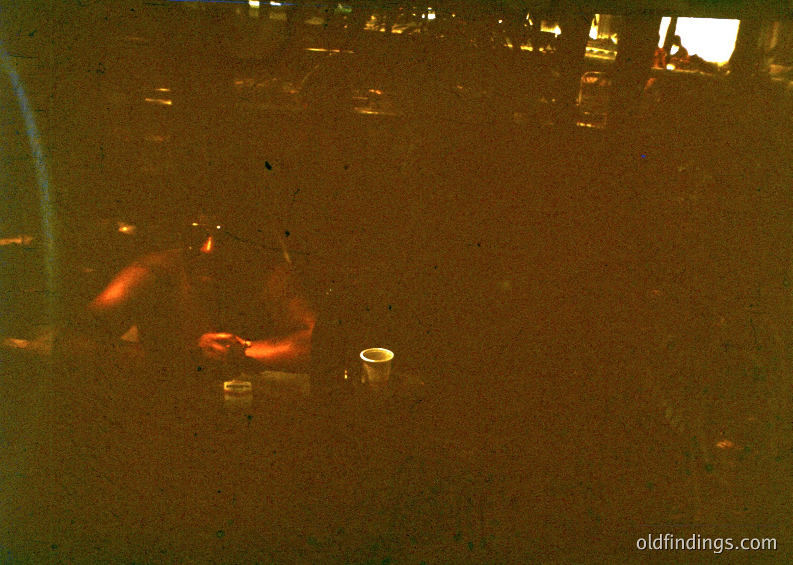 Vintage low-angle shot of a person reclining on a sandy beach, silhouette framed by warm amber tones. Empty glass and cigarette pack suggest mid-20th-century leisure. Coastal setting with blurred background of dunes or vegetation.