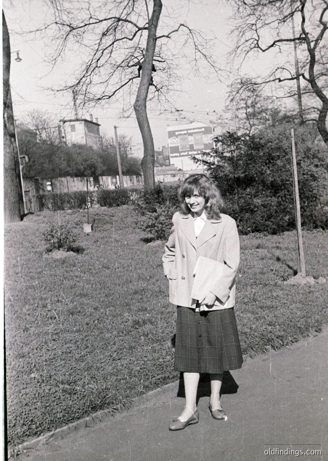 Young woman in 1960s urban park attire—beige coat, pleated skirt, and white gloves—holding a small bag. Leaves suggest late autumn/winter. Mid-rise buildings and bare trees in background indicate a European cityscape.