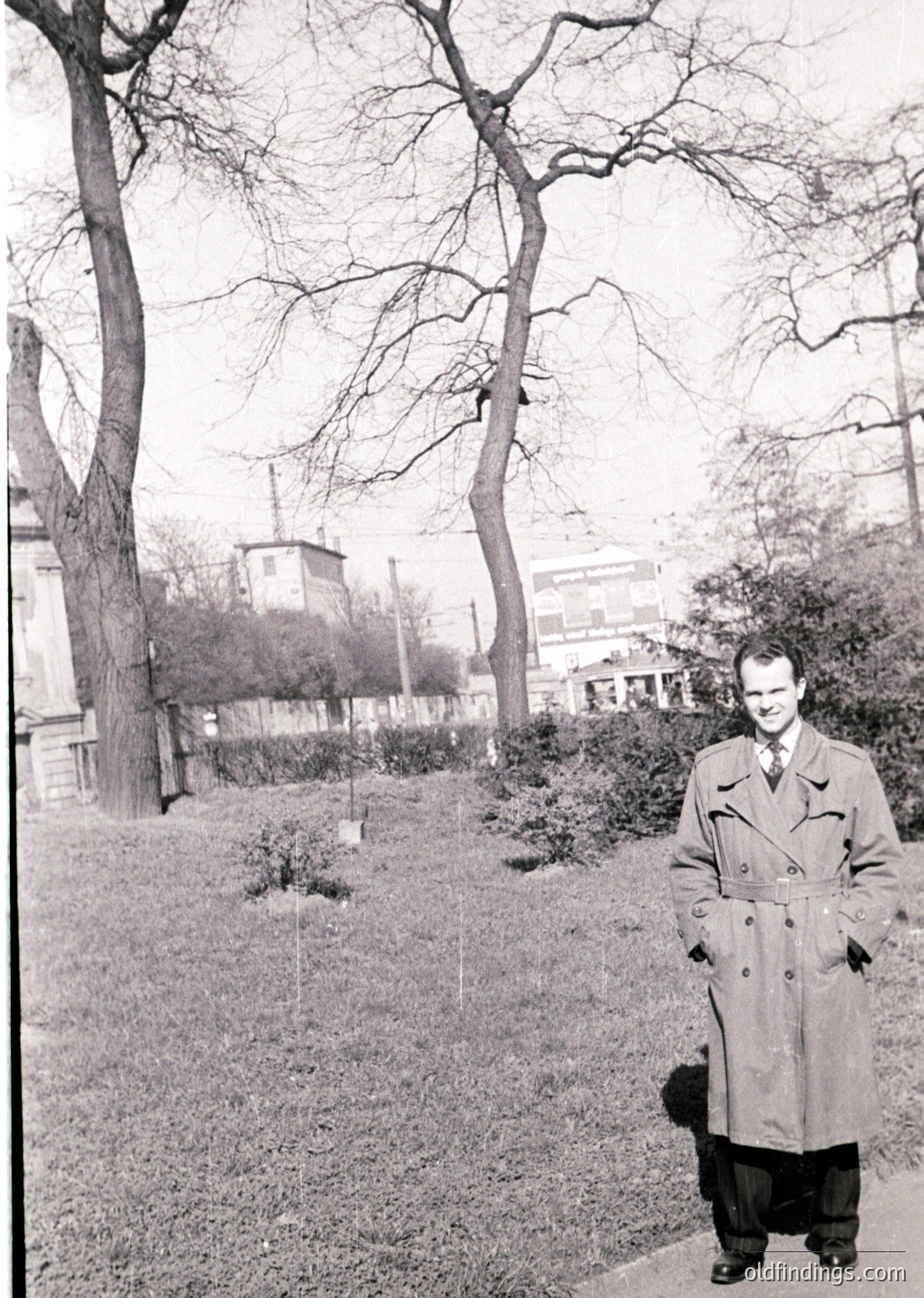 Mid-century man in a long overcoat poses outdoors in a park-like setting, likely 1950s–1960s. Bare trees and a distant industrial tower frame the scene, suggesting urban greenery. Classic streetwear and monochrome film evoke vintage photography.