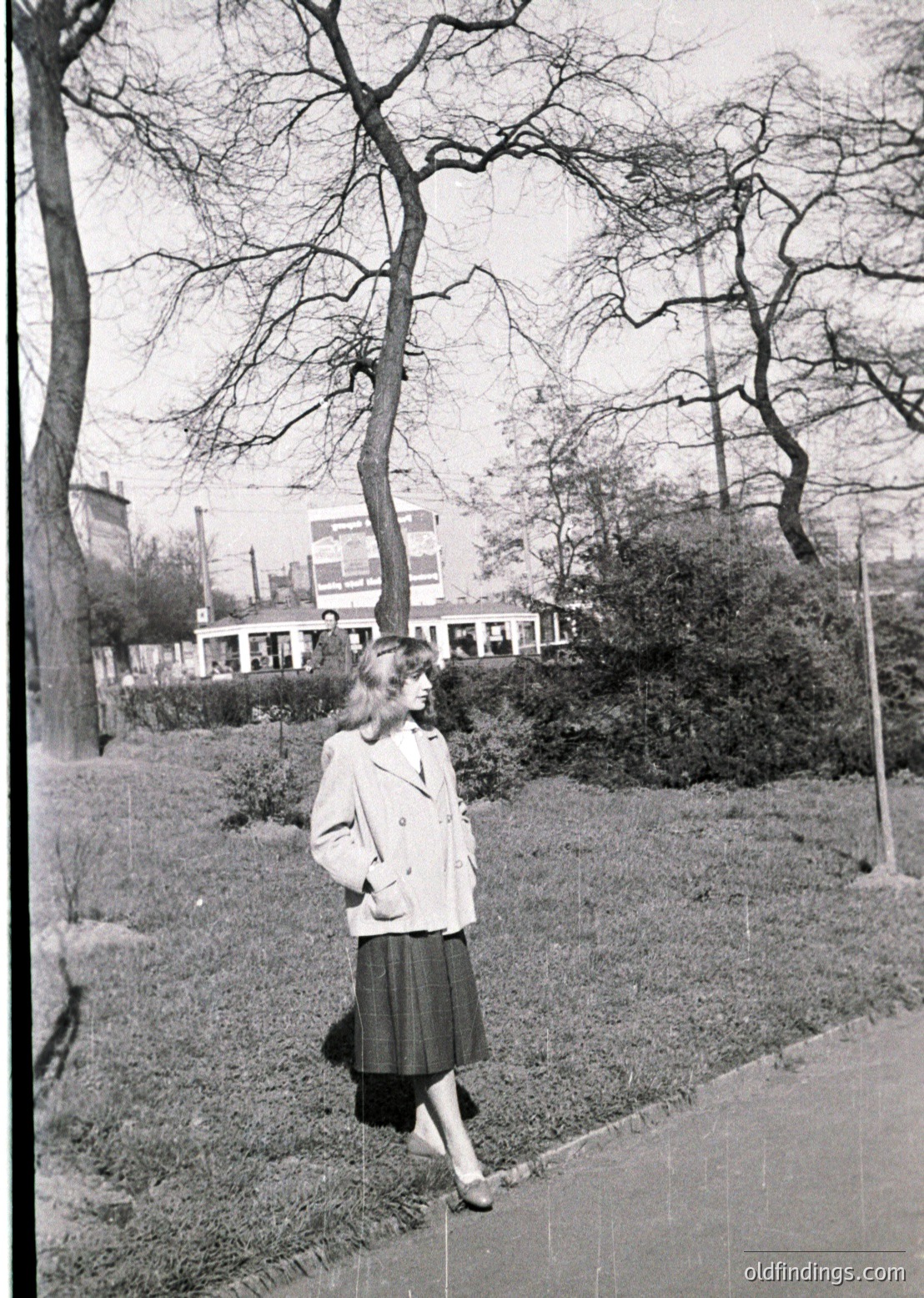 Mid-century woman in a light blazer and pencil skirt strolls along a paved path in a park, flanked by bare winter trees. Urban buildings and a tram line visible in background, suggesting European cityscape.