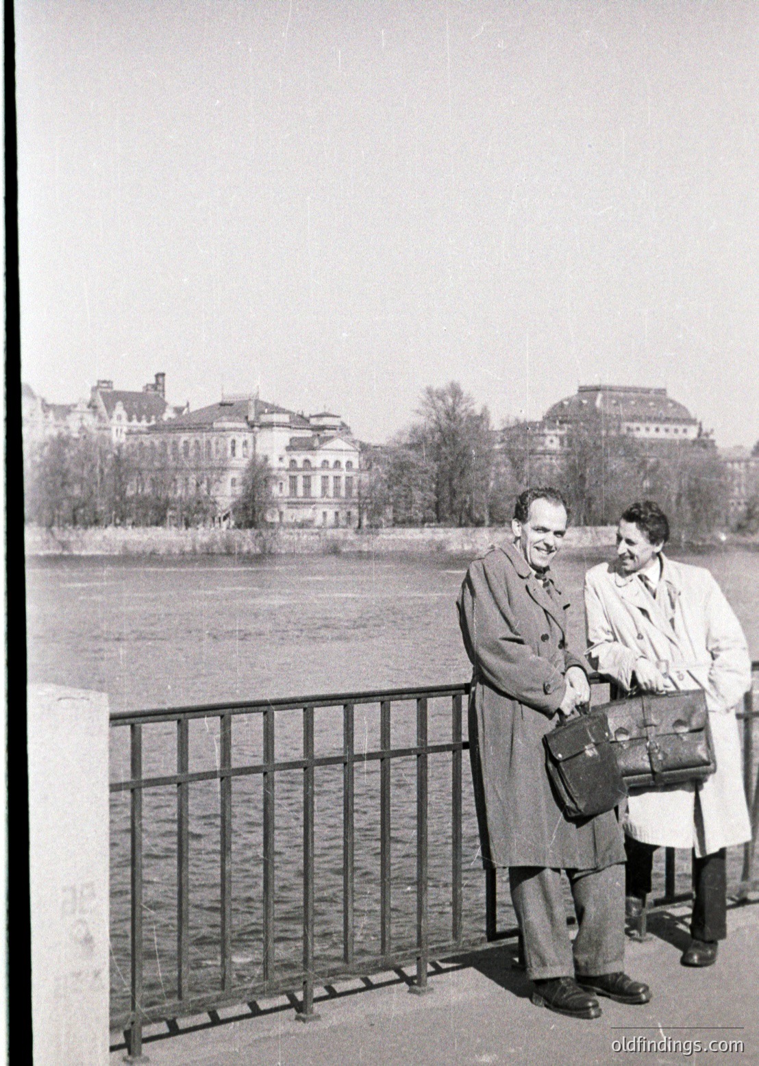 Two men in mid-20th century attire pose by a riverbank railing, one holding a briefcase. Behind them, grand neoclassical buildings with mansard roofs and symmetrical windows line the waterfront. Overcast skies suggest early-to-mid 1900s European urban setting.