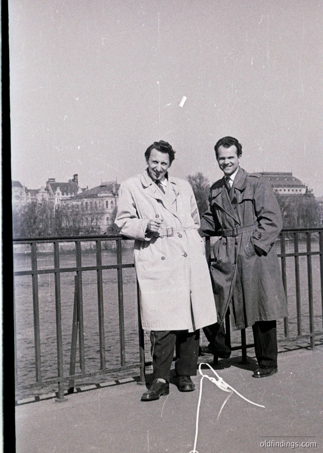 Mid-20th century black-and-white photo of two men in overcoats posing on a bridge railing by a river, likely 1950s–1960s. Urban backdrop features classical architecture with domes and spires, suggesting Eastern European setting. One man holds a small object, possibly a cigarette case or compact.