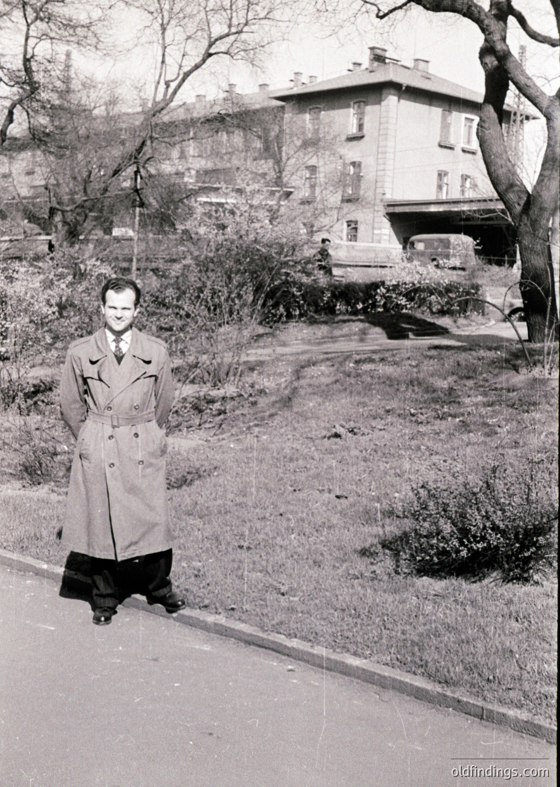 Mid-century man in a belted trench coat poses on a tree-lined urban sidewalk, 1950s–60s. Multi-story residential building with classic brickwork and parked vintage car in background. Black-and-white street photography captures mid-20th century urban lifestyle.