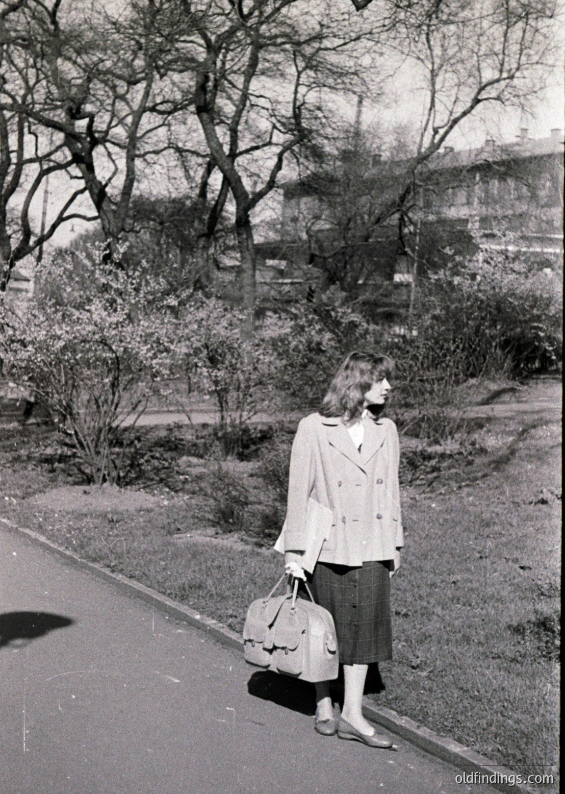 Mid-century street portrait: Woman in a structured blazer, pleated skirt, and knee-high socks stands on a paved path, holding a vintage suitcase. Leafless trees and urban buildings suggest late autumn/winter.
