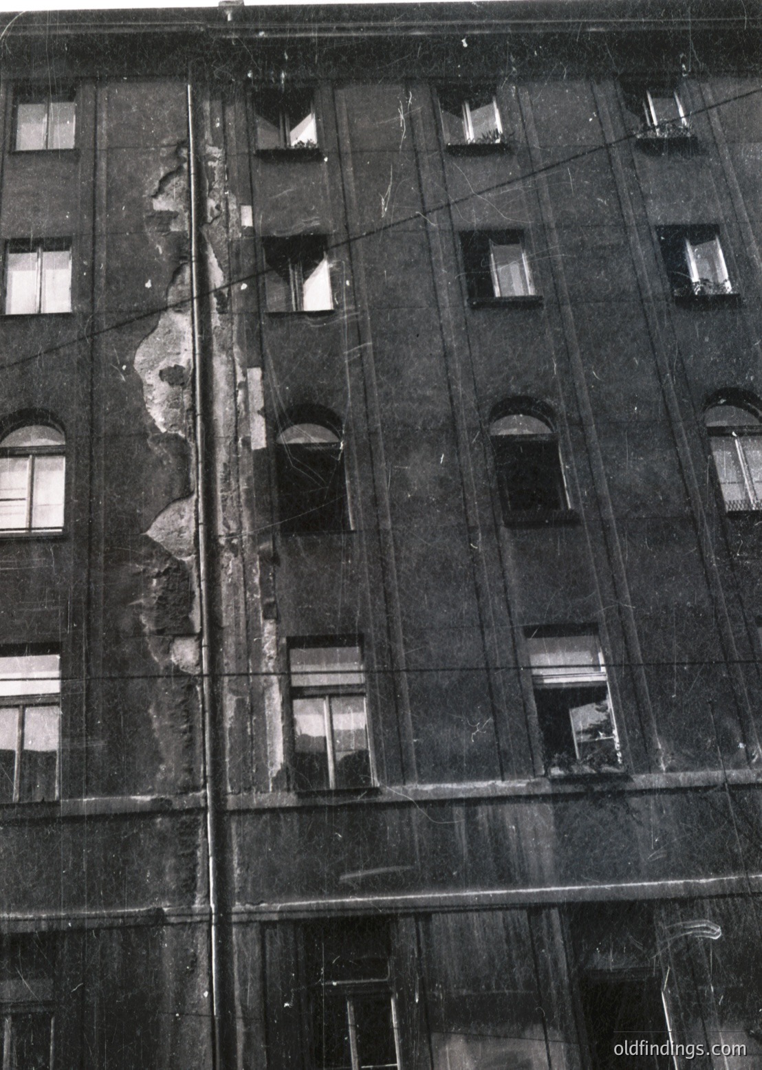 Mid-century Soviet-era concrete apartment block façade, showing structural damage and exposed rebar. Symmetrical arched windows and vertical utility lines. Likely Eastern Europe, 1960s–1980s.
