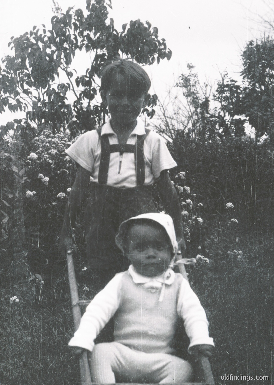 Vintage black-and-white portrait of a young boy standing, holding an infant seated on a bench in an outdoor setting with dense foliage. Boy wears suspenders and a short-sleeved shirt; infant dressed in a long-sleeved dress with a bonnet. Likely mid-20th century, possibly 1940s–1950s.