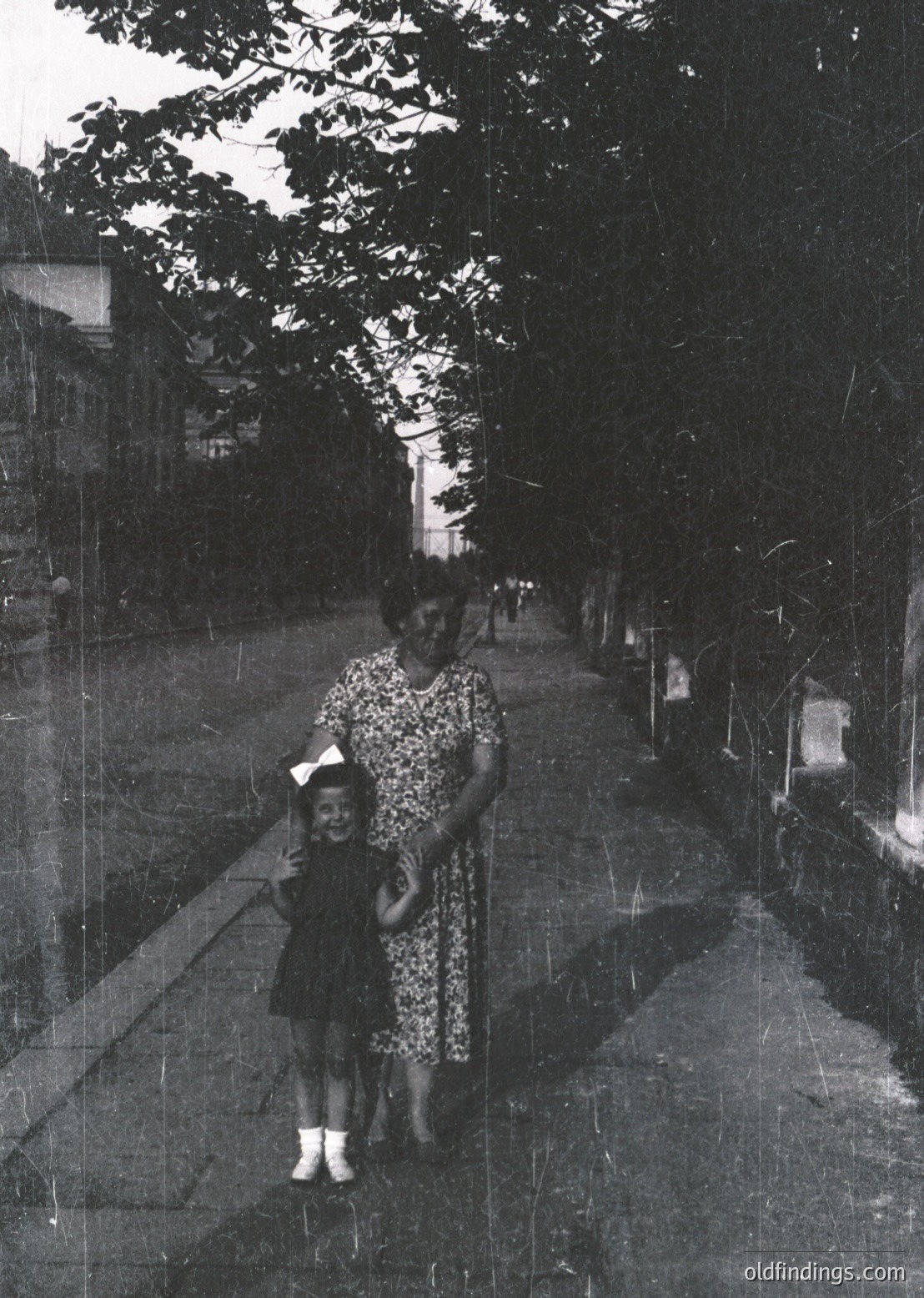 A mid-20th century black-and-white photo of a woman and child walking on a tree-lined path, likely 1950s–1960s. The woman wears a floral dress with a belted waist, while the child dons a hat and a dark dress with white socks. The paved pathway and mature trees suggest an urban or suburban park setting.