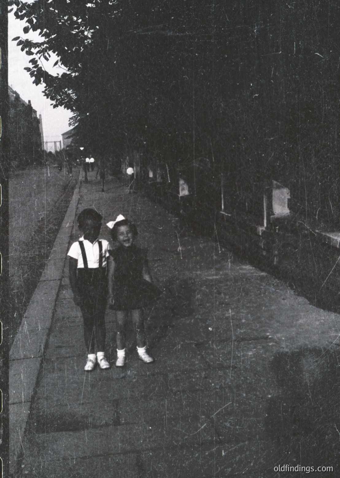 Two children pose on a wide, tree-lined concrete pathway, likely mid-20th century. Girl in a knee-length dress with white socks and shoes; boy in overalls with white shoes. Pathway flanked by low stone walls and sparse lighting. Vintage black-and-white film grain evident.