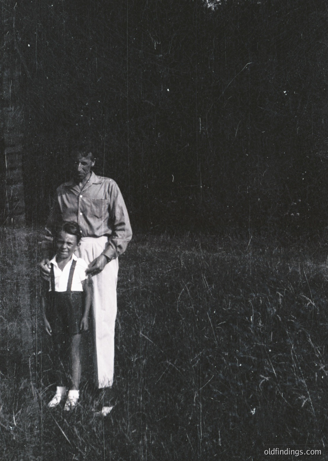 A mid-20th century black-and-white portrait of an adult holding a young child outdoors in overgrown grass. The adult wears a button-up shirt and light-colored pants, while the child dons a short-sleeve shirt and dark pants. The setting appears rural or semi-rural, with a dark, textured wall in the background.