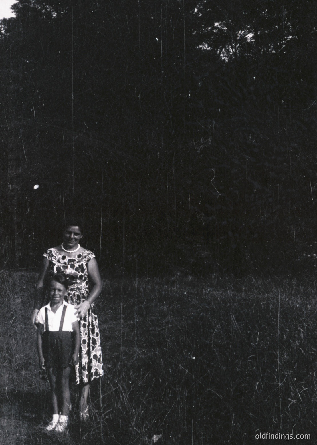 Black-and-white portrait of a woman and child in a grassy outdoor setting, likely 1950s–1960s. Woman wears a leopard-print dress with white socks and sandals; child in short-sleeve shirt and shorts. Dense foliage and shadows dominate the background.