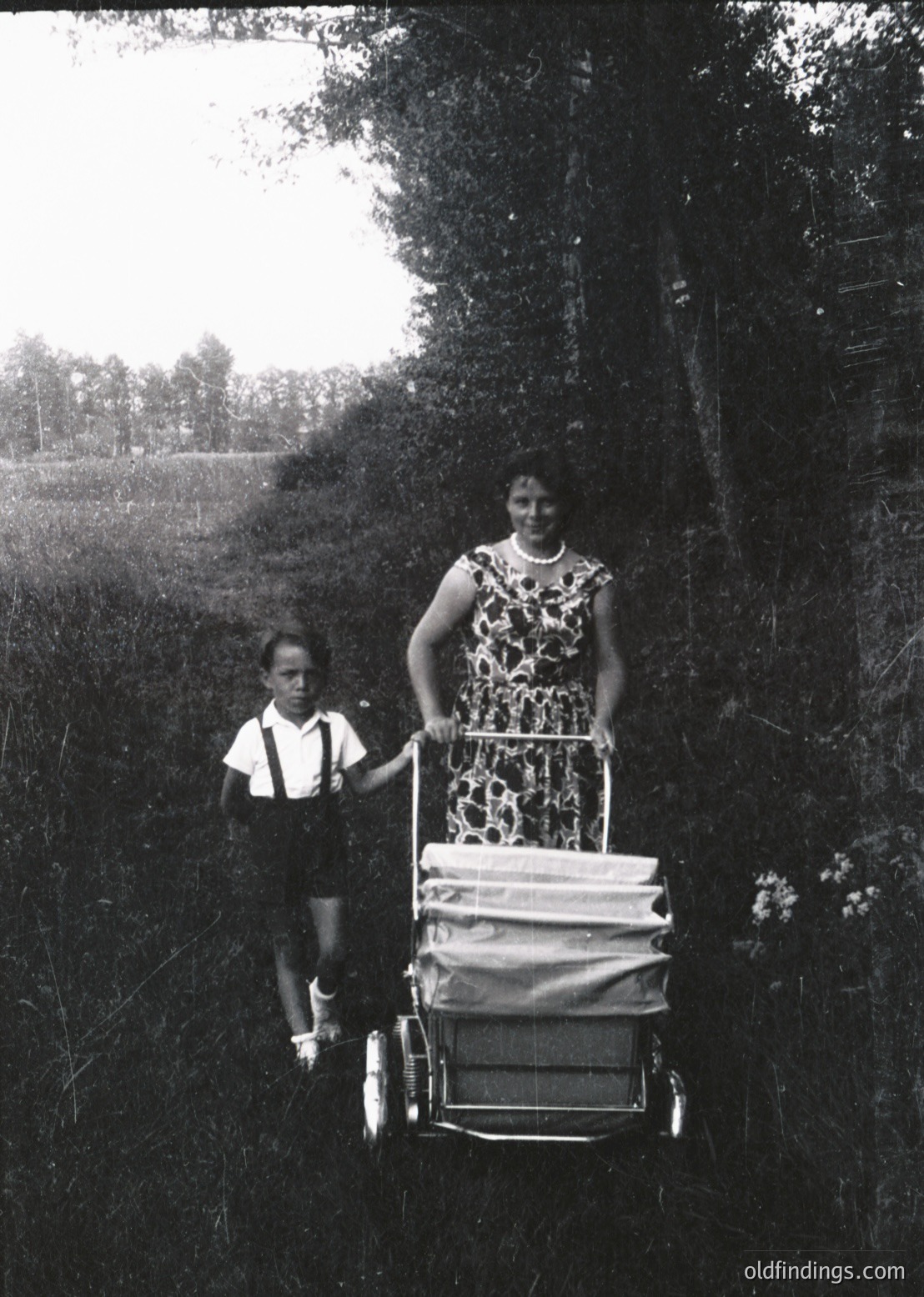 Black-and-white photo of a woman pushing a child in a wheeled cart along a rural path, likely mid-20th century. Floral dress and boy’s short-sleeved shirt suggest 1950s–1960s fashion. Dense greenery and open field in background indicate countryside setting. Ideal for vintage lifestyle, historical research, or nostalgic design references.