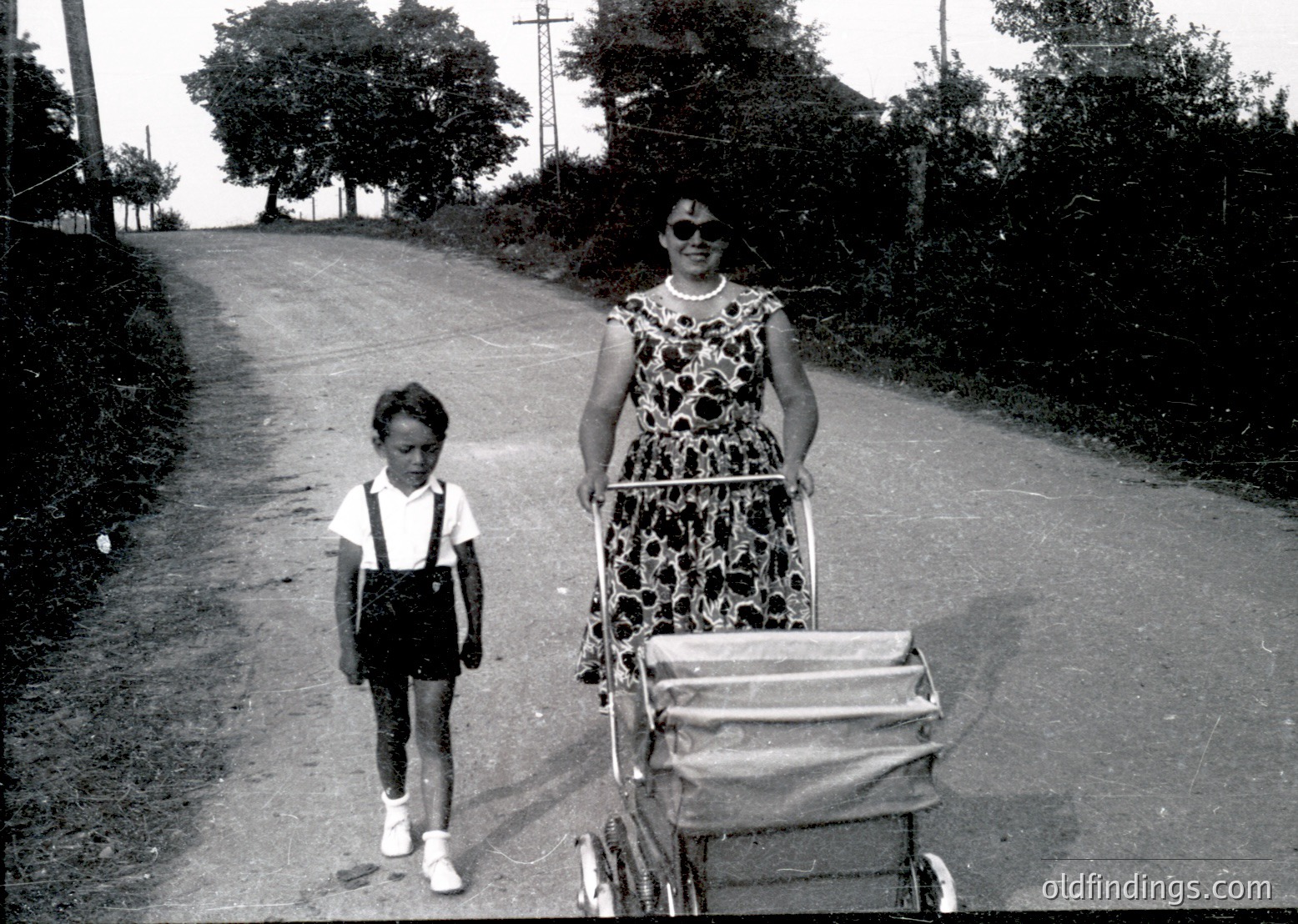 Black-and-white street scene featuring a woman in a floral dress and sunglasses pushing a vintage pram, accompanied by a young boy in suspenders. Rural road flanked by dense greenery and mature trees, suggesting mid-20th century.