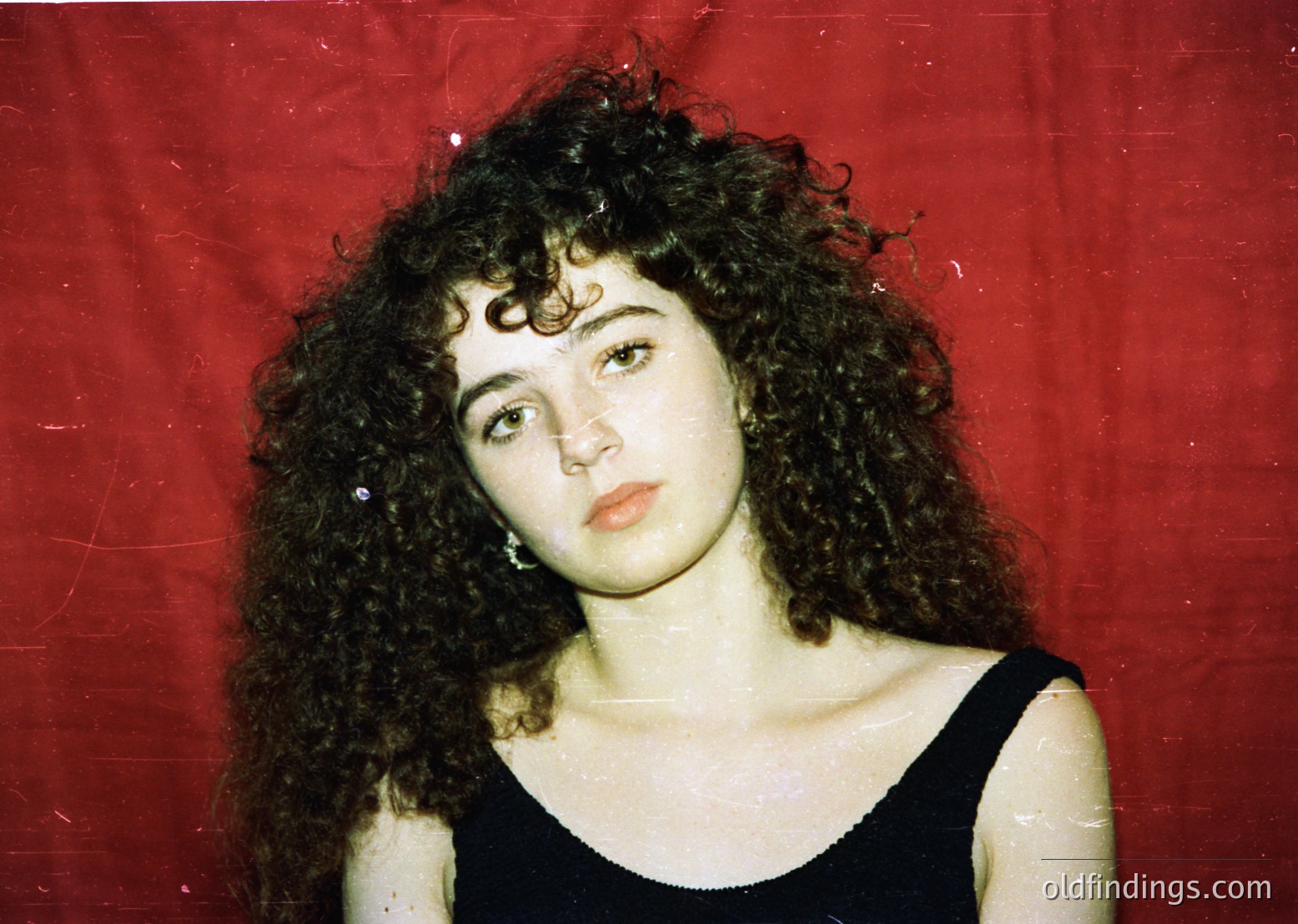 Portrait of a woman with voluminous curly hair, shot against a faded red backdrop. Her expression is neutral, wearing a sleeveless black top. Likely 1980s–1990s fashion, evoking retro or vintage aesthetic.