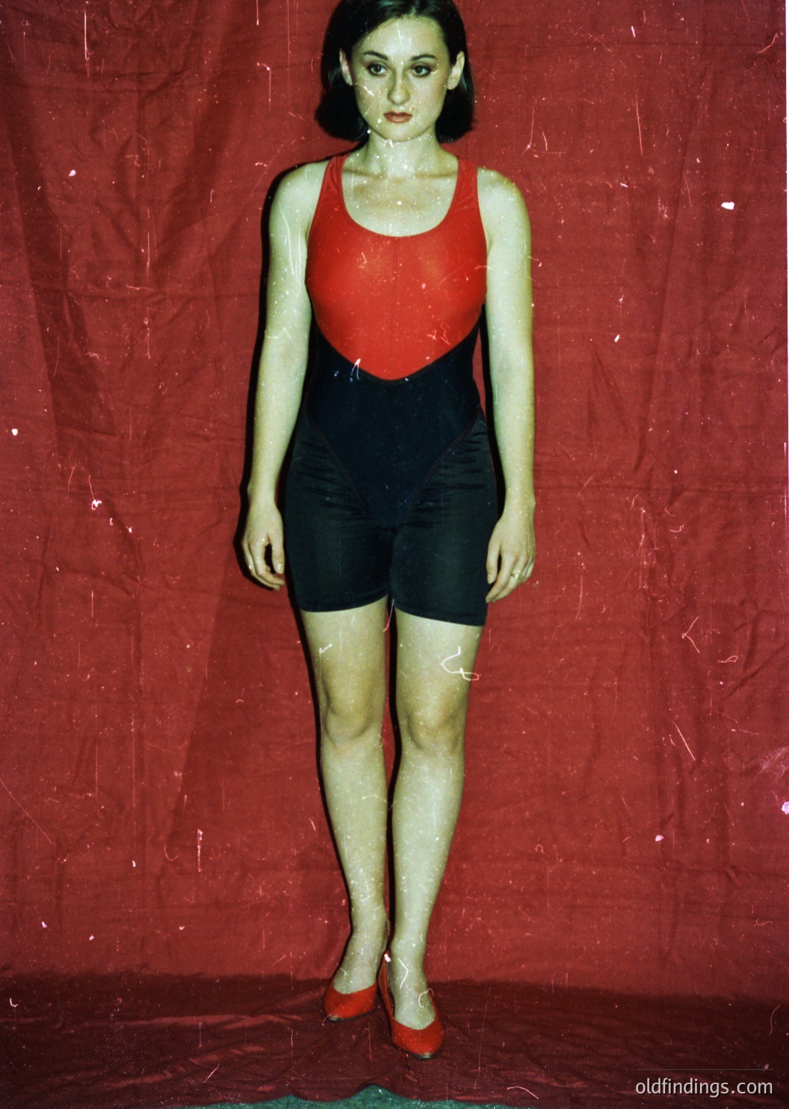 Vintage mannequin in bold 1960s-style swimsuit: red top with black paneling, black shorts, and red shoes. Stylized pose on red backdrop suggests promotional or fashion context.