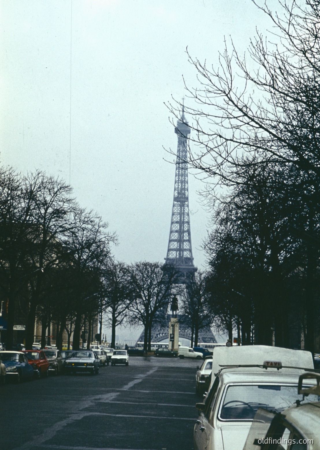 Classic Eiffel Tower framed by bare winter trees, Paris . Narrow street lined with vintage cars and parked vehicles, evoking mid-century urban life. Overcast sky enhances iconic silhouette.