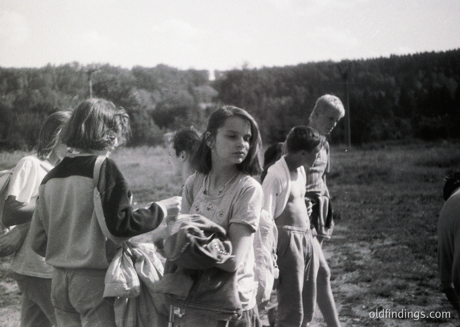 Group of children in outdoor setting, mid-20th century. Central figure holds a basket, others gather around. Casual clothing suggests informal gathering or school outing. Wooded background indicates rural or park location.