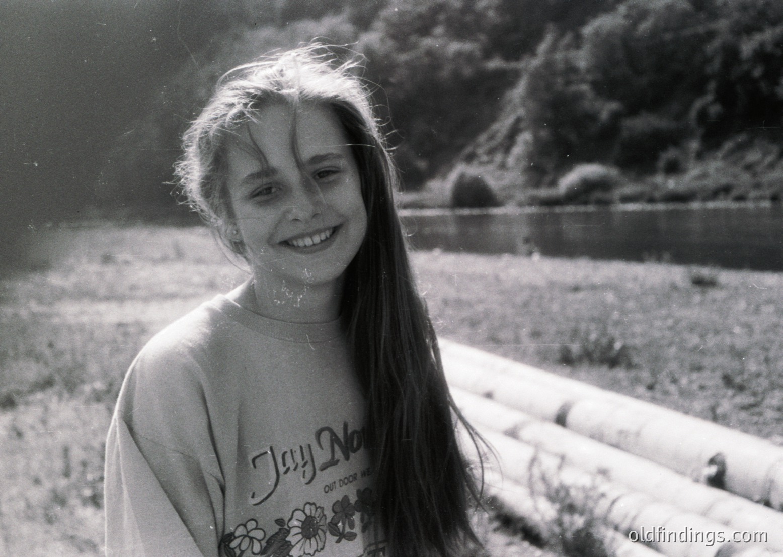 Black-and-white candid of a smiling child outdoors, wet hair framing face with sunlight rays creating lens flare. "Jay Na" graphic shirt suggests 1970s–1980s era. Natural setting with wooden logs and water body in background.