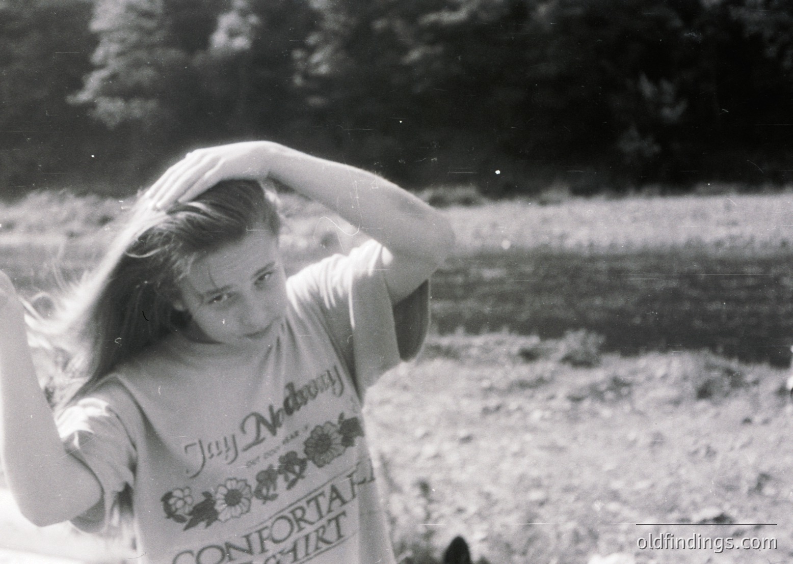 Vintage black-and-white photo of a young woman in a "Jay McDermott Comfort" t-shirt, outdoors near a body of water. Her hair is windblown, suggesting a breezy setting. The image evokes 1970s casual fashion and outdoor recreation.