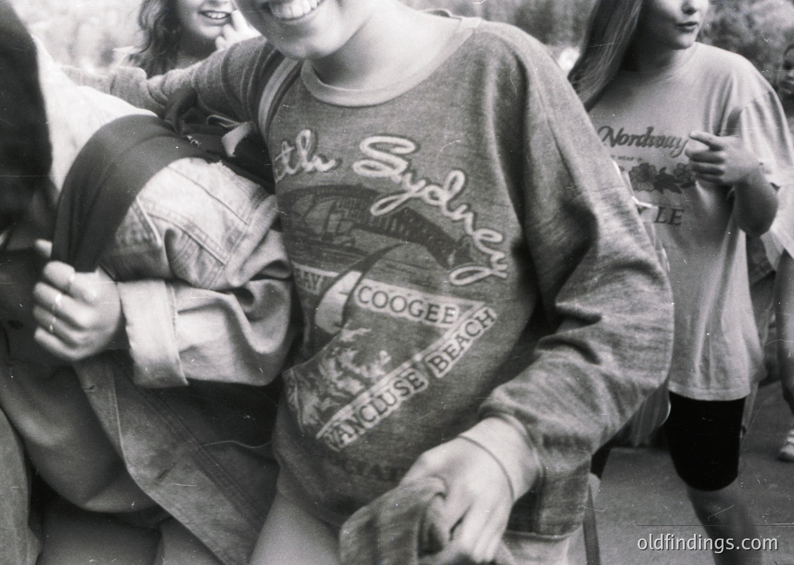 Black-and-white photo of three young women hugging outdoors, wearing 1970s-style sweatshirts with Sydney beach logos ("Coogee Beach" & "Wendy’s"). Casual, candid moment captures 1970s Australian youth culture.