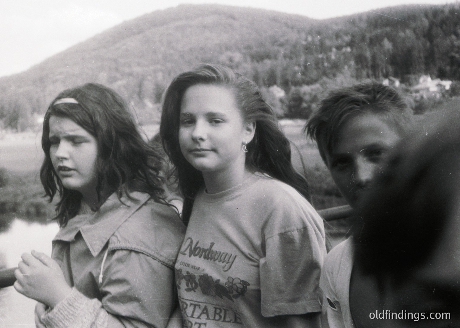 Black-and-white portrait of three young women in outdoor setting, likely 1970s. Center figure wears a "Nordway Portable" t-shirt, suggesting American or European travel influence. Forest and river backdrop hints at hiking or nature trip. Candid expressions and casual attire evoke youth culture of the era.