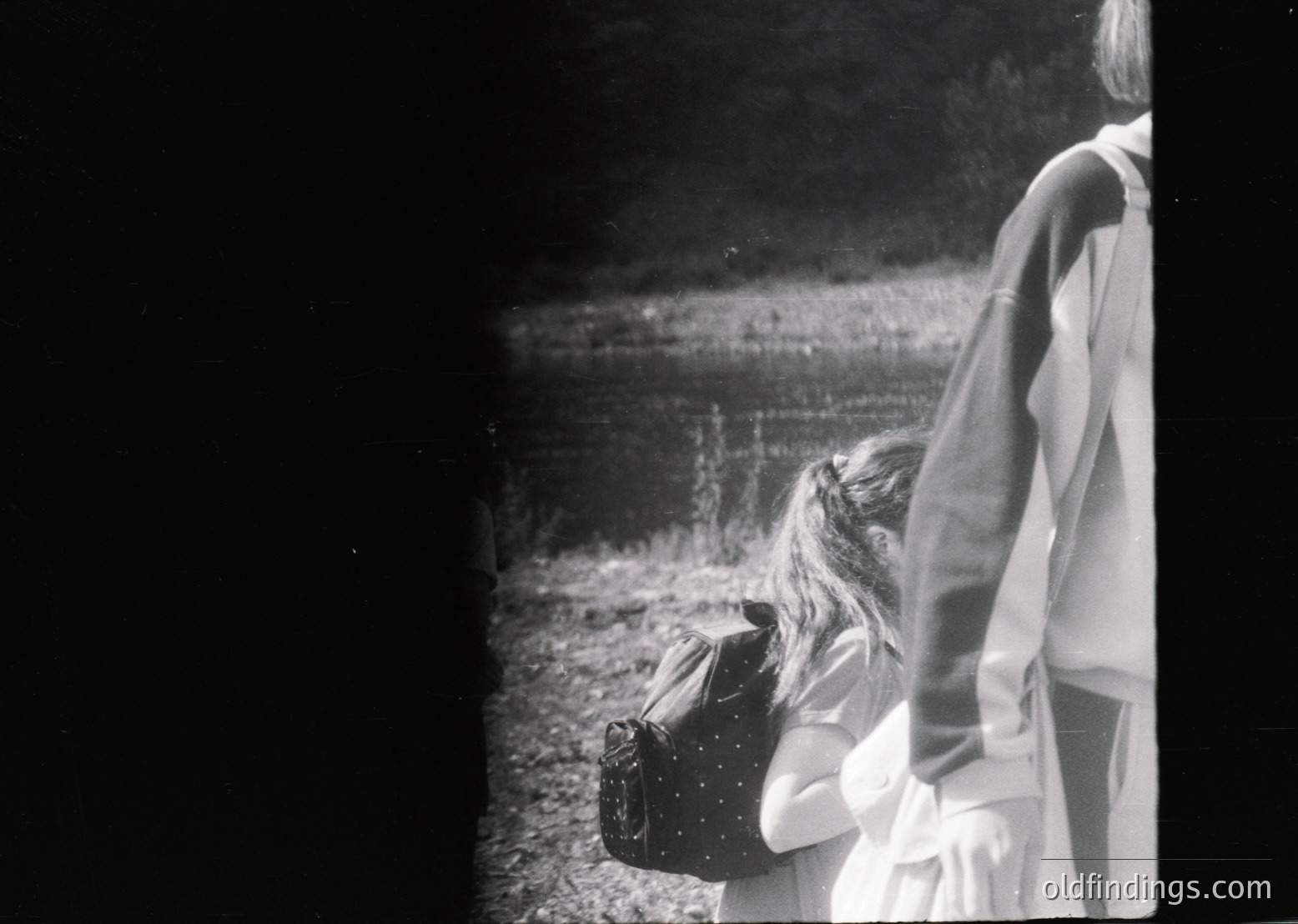 Vintage black-and-white photo of two children standing near a body of water, one holding a vintage suitcase. The scene suggests a mid-20th-century travel or family outing. The blurred background hints at motion or depth.