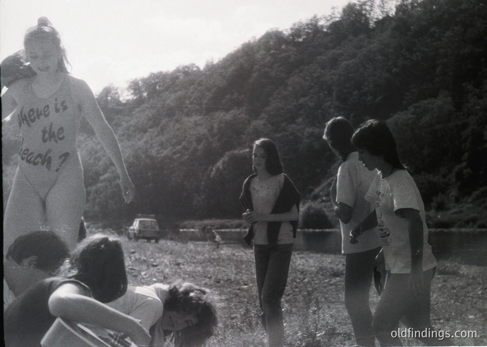 Black-and-white protest scene: A large inflatable cutout of a person holding a sign reading *"Where is the beach?"* draws attention in a forested, riverside area. Group of people, likely 1970s, engage with the prop.