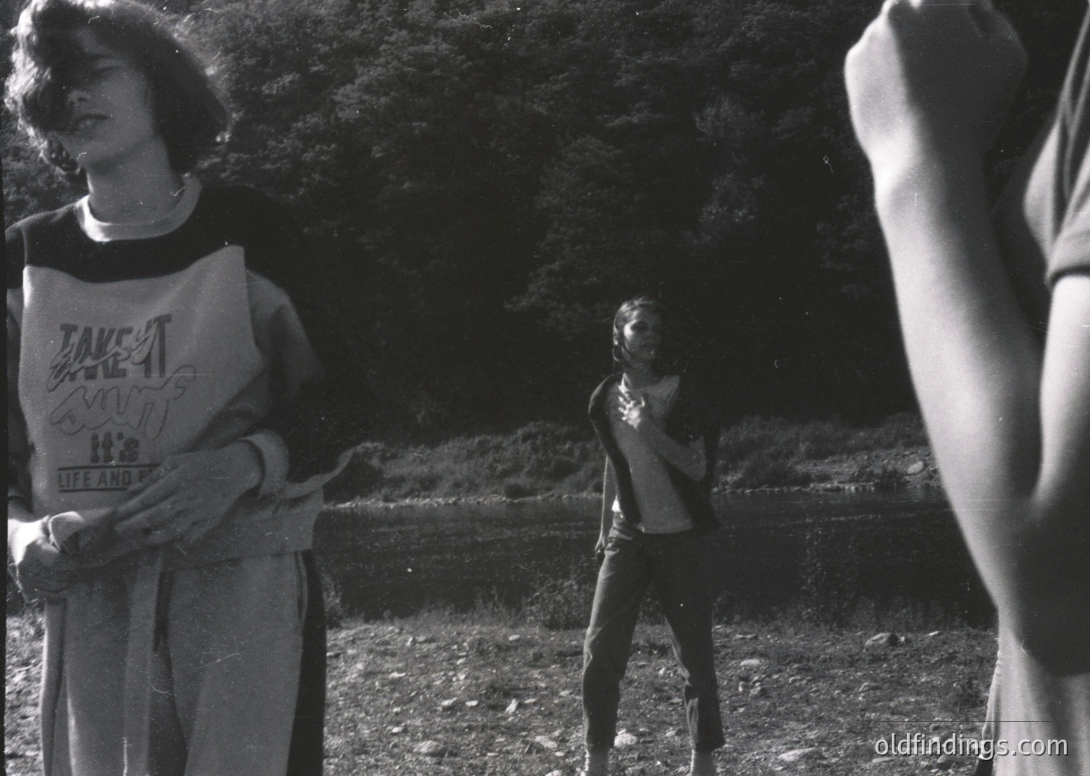 Black-and-white candid of three young people in outdoor setting, likely 1970s–1980s. Foreground: person in loose-fitting sweatshirt with "Lake Atitlán" text and "Off the Grid" slogan, holding a cigarette. Midground: another individual in a sleeveless top, arms crossed. Background: dense foliage and blurred motion. Urban/suburban park vibe.
