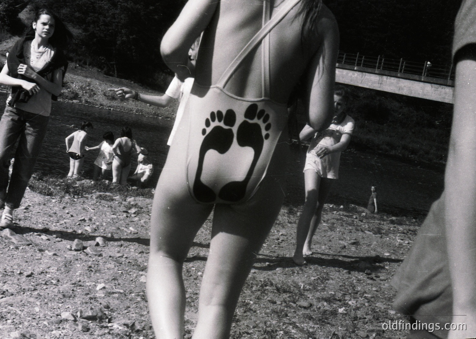 Black-and-white photo capturing 1970s beachgoers in swimwear with playful paw-print designs. Group of people lounging on rocky terrain near water, suggesting a natural, informal seaside gathering. Casual attire reflects mid-century beach culture.