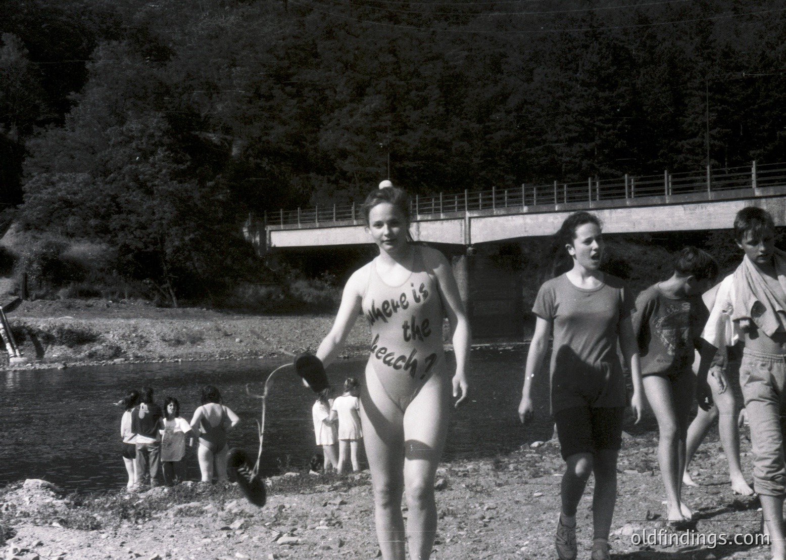 Vintage black-and-white photo of a group by a riverside in the 1960s–70s. Central figure wears a bikini with handwritten text ("Where is the beach?") and holds a bucket. Concrete bridge and lush greenery frame the scene. Casual summer attire and relaxed poses suggest a social outing or picnic.
