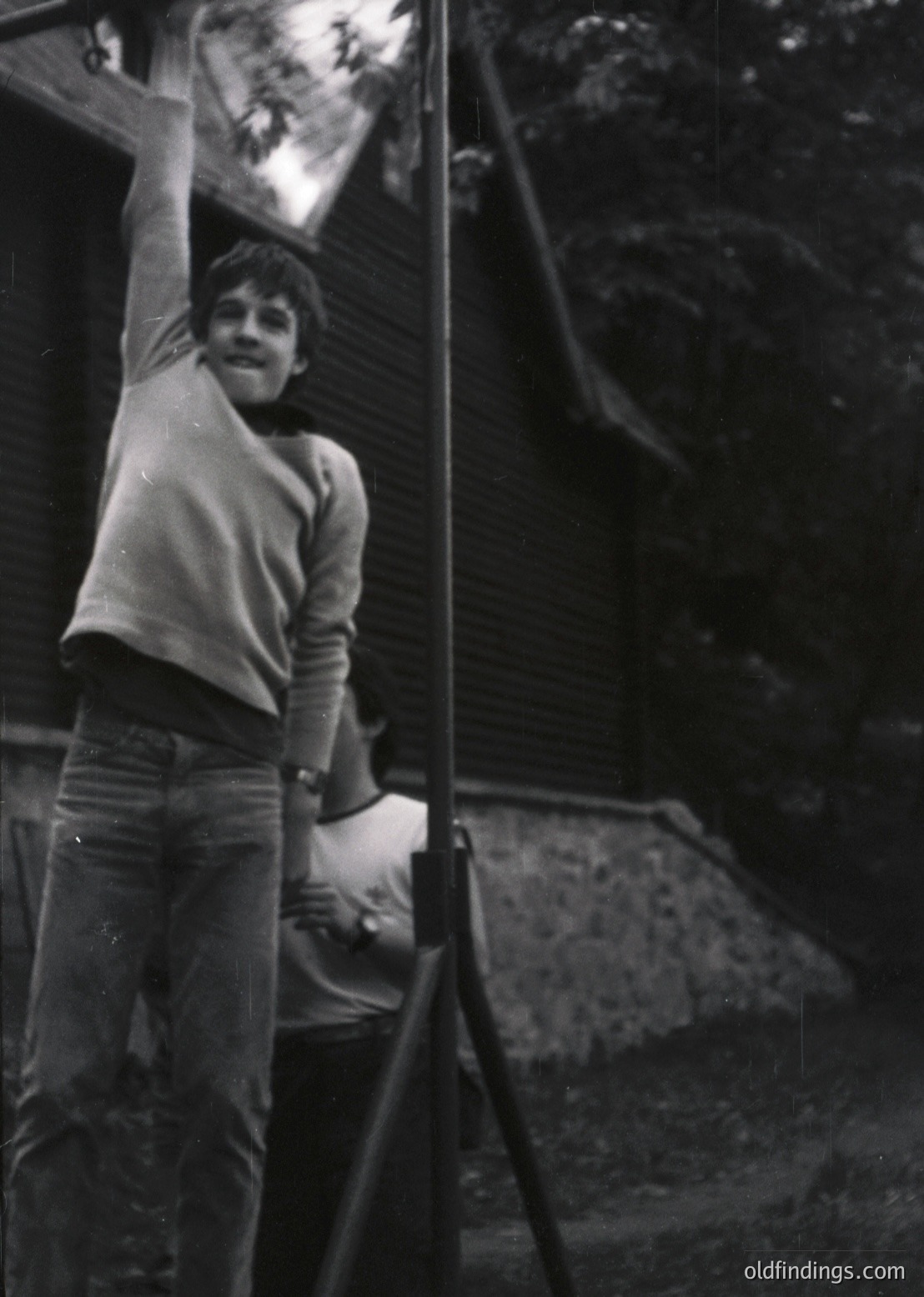 Mid-century youth poses on wooden ladder against dark stone wall, mid-1960s. Casual attire—sweater, jeans—suggests Western European or North American setting. Playful expression and relaxed posture evoke carefree era. Ideal for vintage lifestyle, historical research, or nostalgic design references.