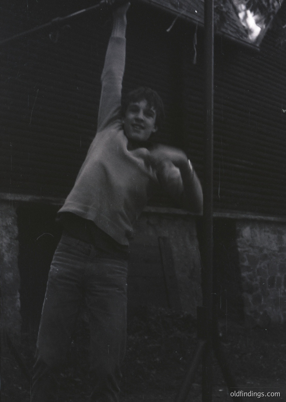 Vintage black-and-white photo of a young boy mid-jump, shirtless with rolled-up pants, against a dark, textured wall. Motion blur suggests playful energy. Likely mid-20th century, possibly 1950s–1960s.