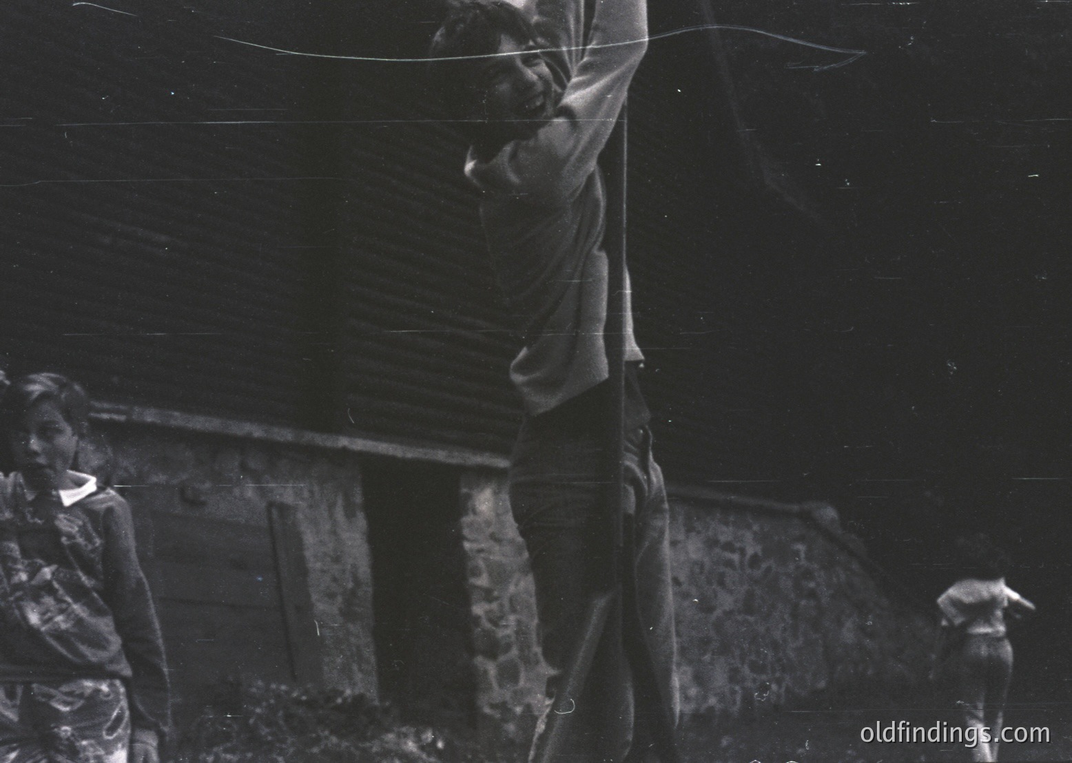 Black-and-white photograph captures three individuals in a rural setting, likely mid-20th century. Central figure stands on a ladder, holding a taut wire or rope against a weathered wooden structure. Another person observes from the left, while a third kneels in the foreground. Rustic wooden house and natural light suggest agricultural or village life.