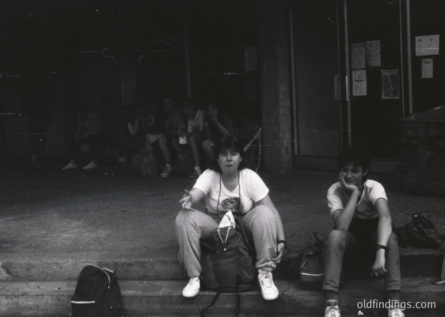 Black-and-white candid shot of two individuals seated on concrete steps in an urban setting, likely 1970s–1980s. The person on the left wears a graphic tee and cargo pants, holding a small object; the right sits with chin resting on hand. Posters and graffiti adorn the brick wall behind them, suggesting a youth culture hub or public space.