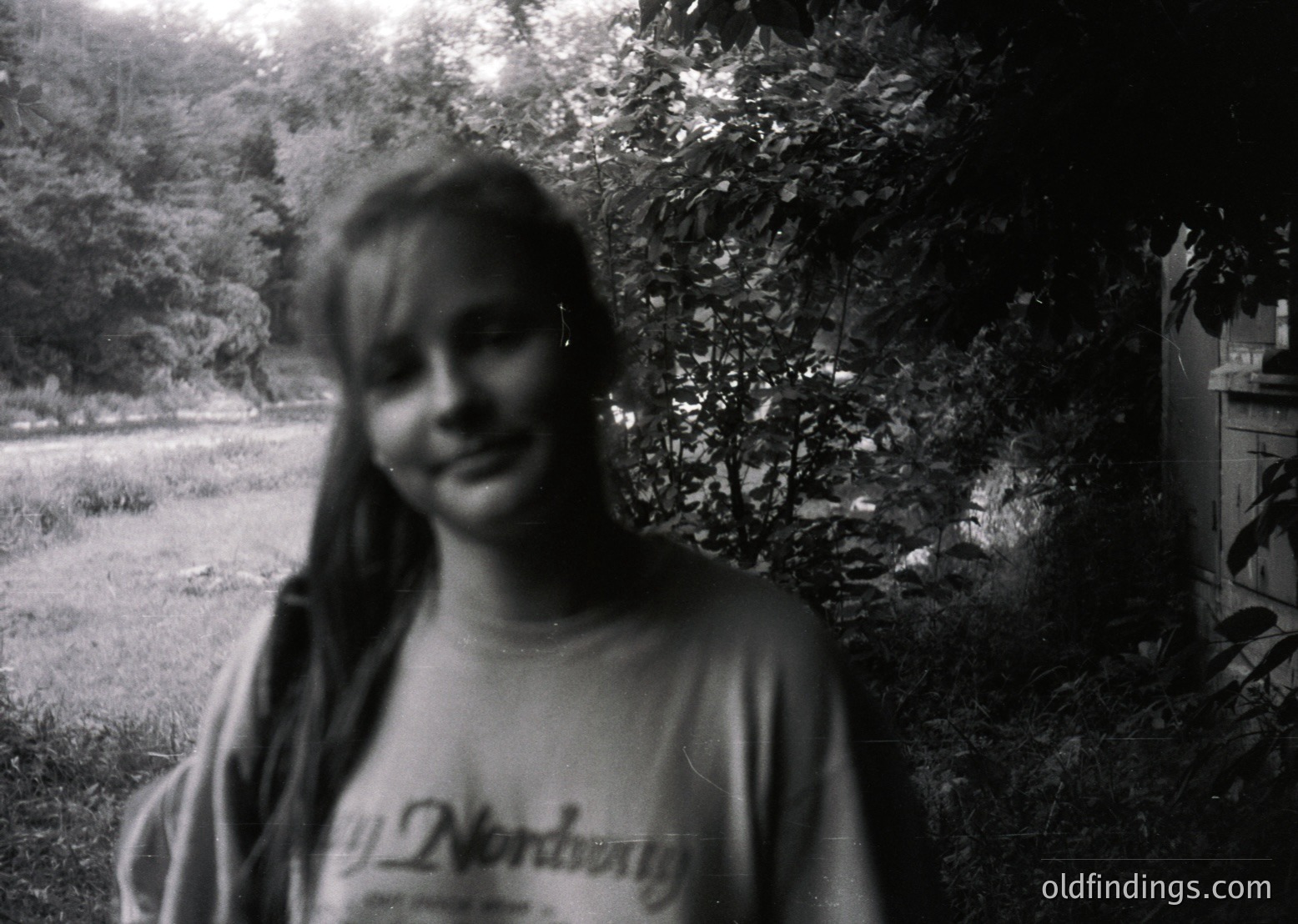 Vintage black-and-white portrait of a smiling individual in a "Nordberg" t-shirt, blurred background with foliage and partial building. Likely mid-20th century outdoor setting.