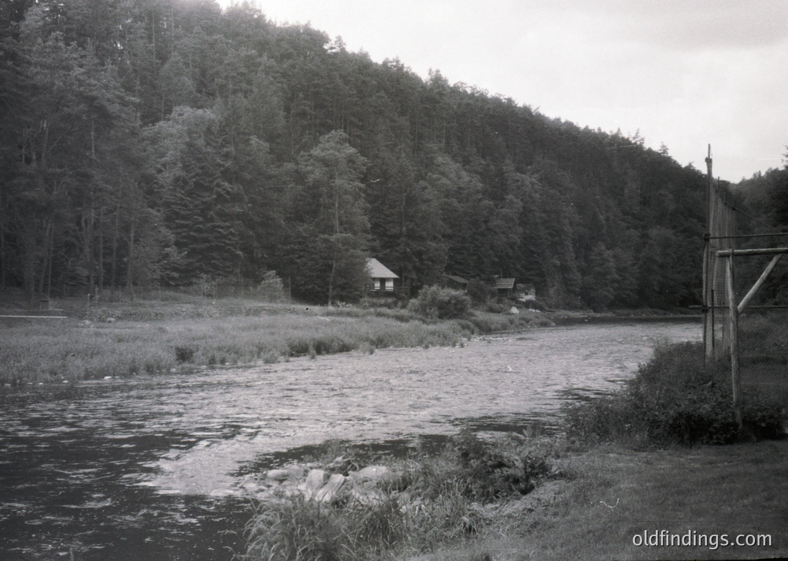 Black-and-white rural scene featuring a shallow river bordered by rocks and reeds, flanked by dense forest. Two modest wooden structures with pitched roofs sit on elevated grassy terrain. Overgrown vegetation and utility poles suggest mid-20th century European countryside.