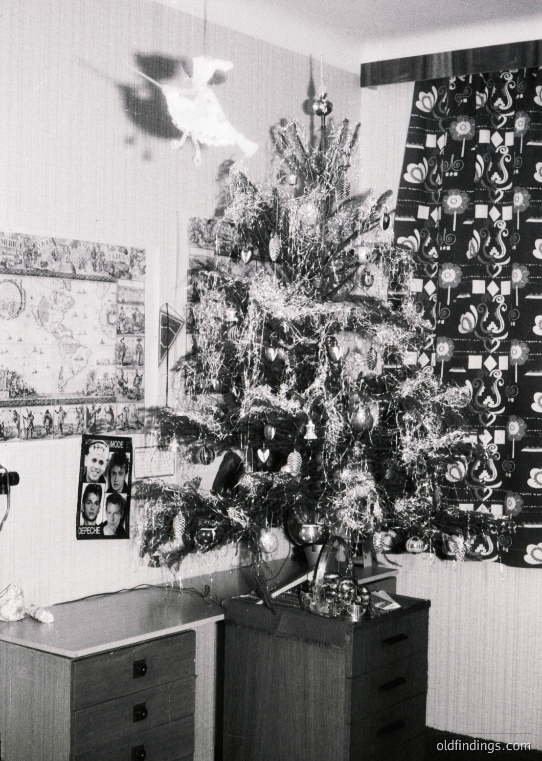 Vintage black-and-white interior featuring a densely adorned Christmas tree with tinsel, geometric ornaments, and a star topper. Mid-century desk with drawers and framed posters of classical art and a portrait. Wall-mounted decorative tiles with floral/symbol motifs. Warm, intimate holiday ambiance.