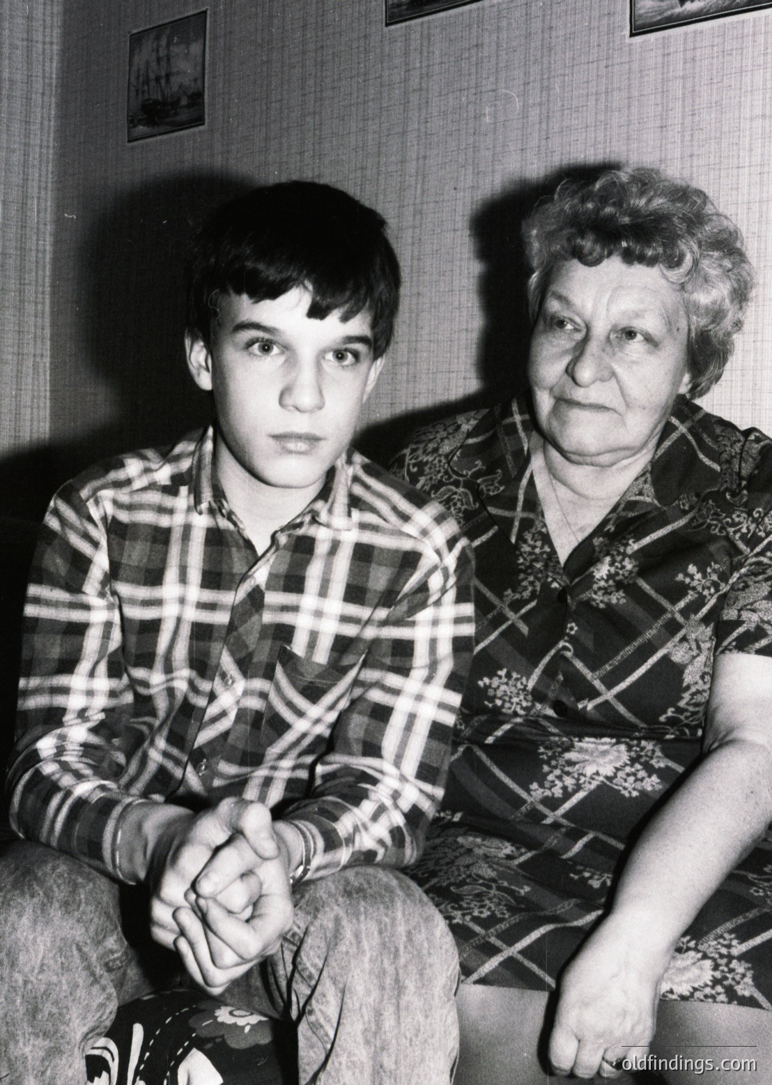 Black-and-white portrait of a young boy and elderly woman in a domestic setting, likely mid-20th century. The boy wears a plaid shirt with rolled sleeves, while the woman’s patterned blouse features geometric embroidery. Their expressions are neutral, framed by a curtained window and framed photos on the wall.