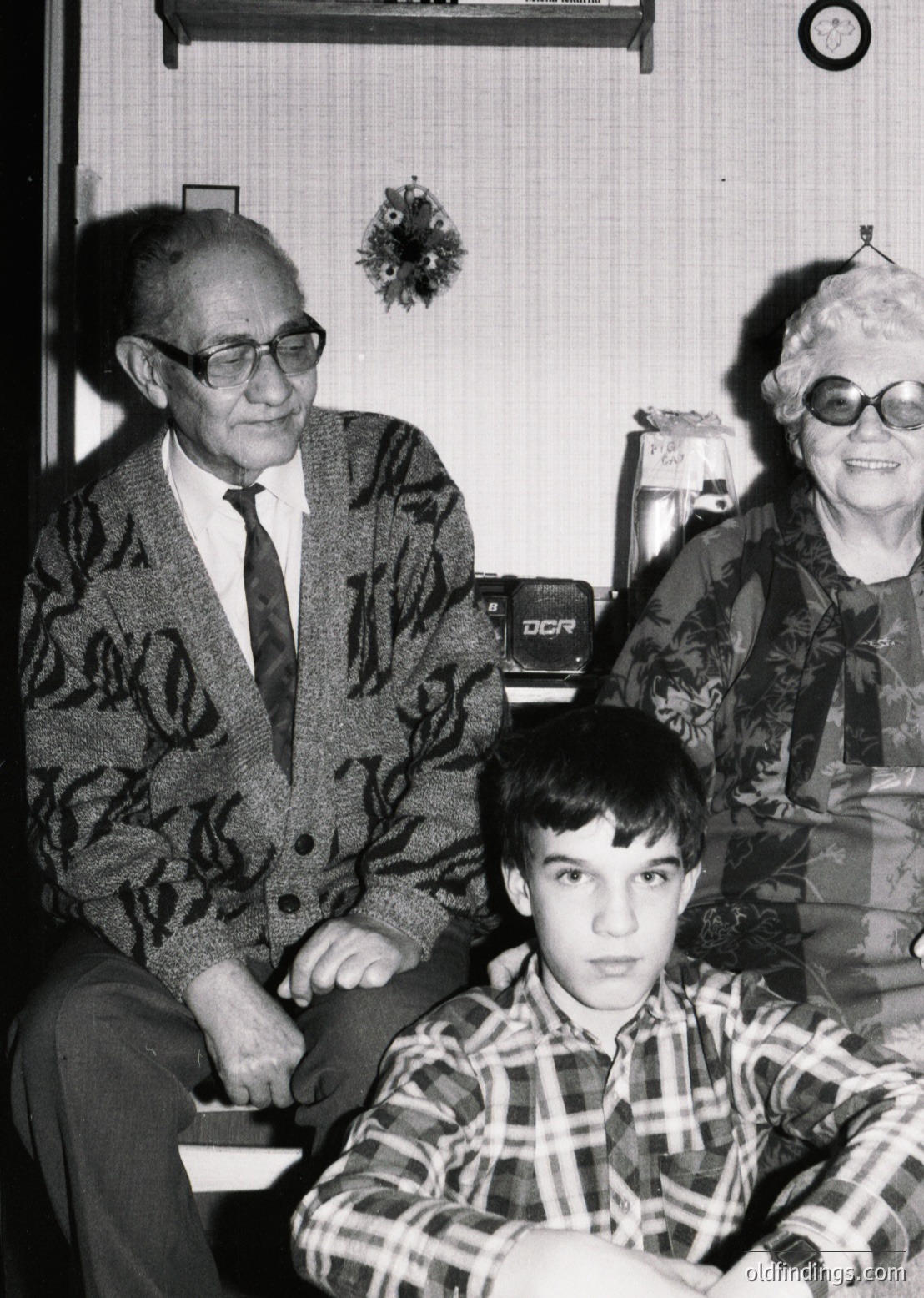 Three-generation family portrait in mid-20th century (likely 1960s–70s) indoors. Elderly man in patterned blazer, woman in floral sweater, and young boy in plaid shirt pose together. Vintage camera (DCR) and decorative wallpaper hint at mid-century home decor.