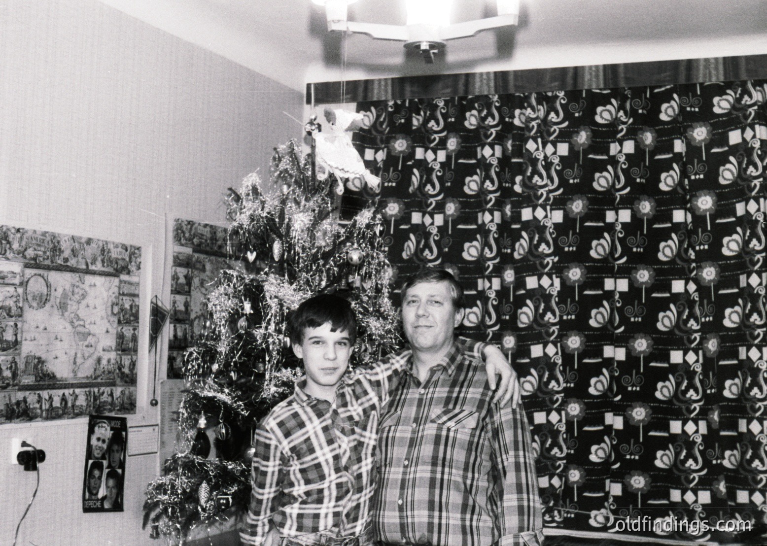 Black-and-white indoor portrait of a man and boy posing beside a decorated Christmas tree and wall adorned with geometric wallpaper and framed photos. Mid-20th century Soviet-era interior, likely 1960s–1970s.