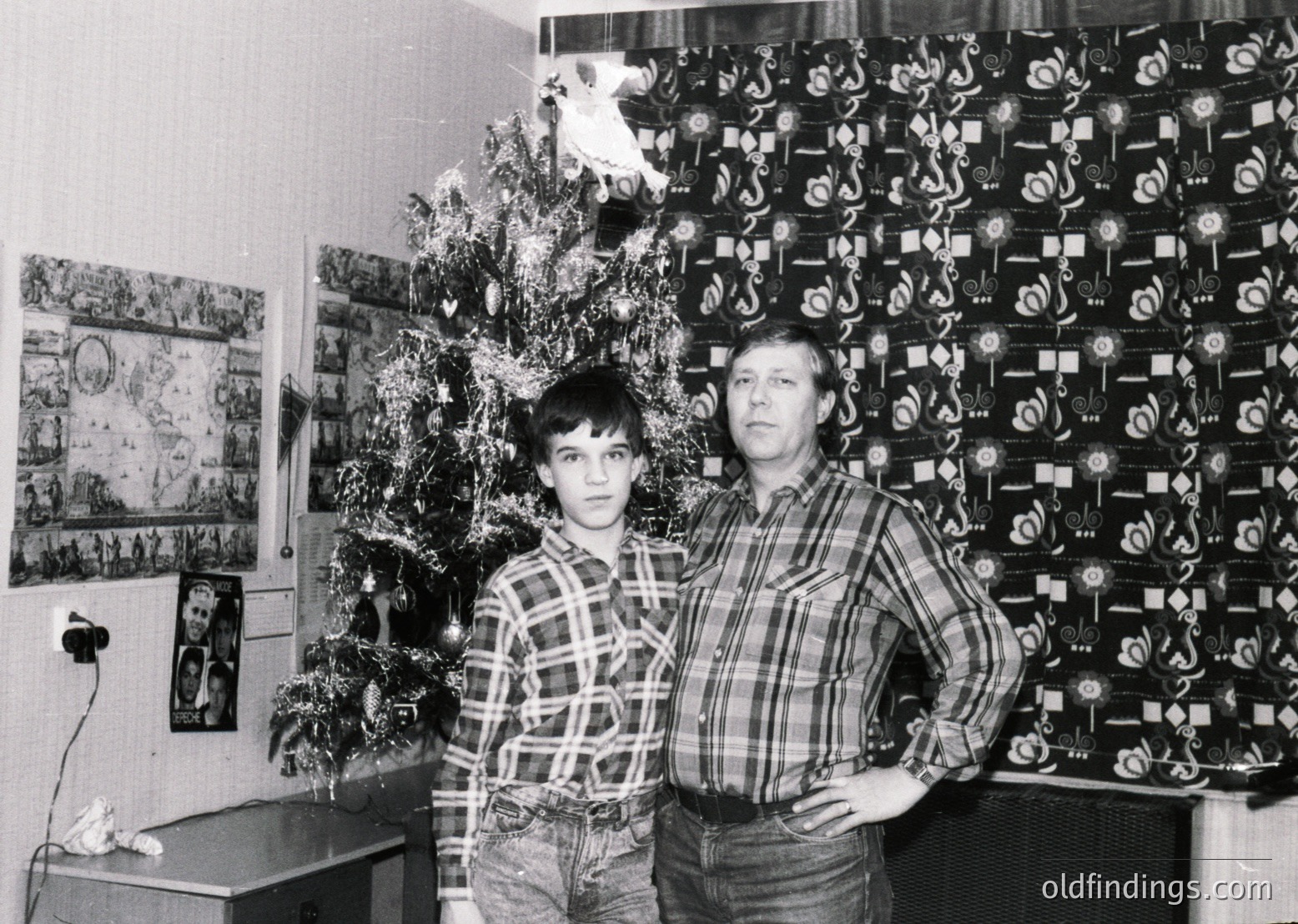 A black-and-white portrait of a man and boy in a Soviet-era home, likely 1970s–1980s. The man wears a plaid shirt, the boy a checkered one; both pose beside a decorated Christmas tree with geometric wallpaper and framed posters. Functional, modest interior reflects mid-century Soviet domestic life.