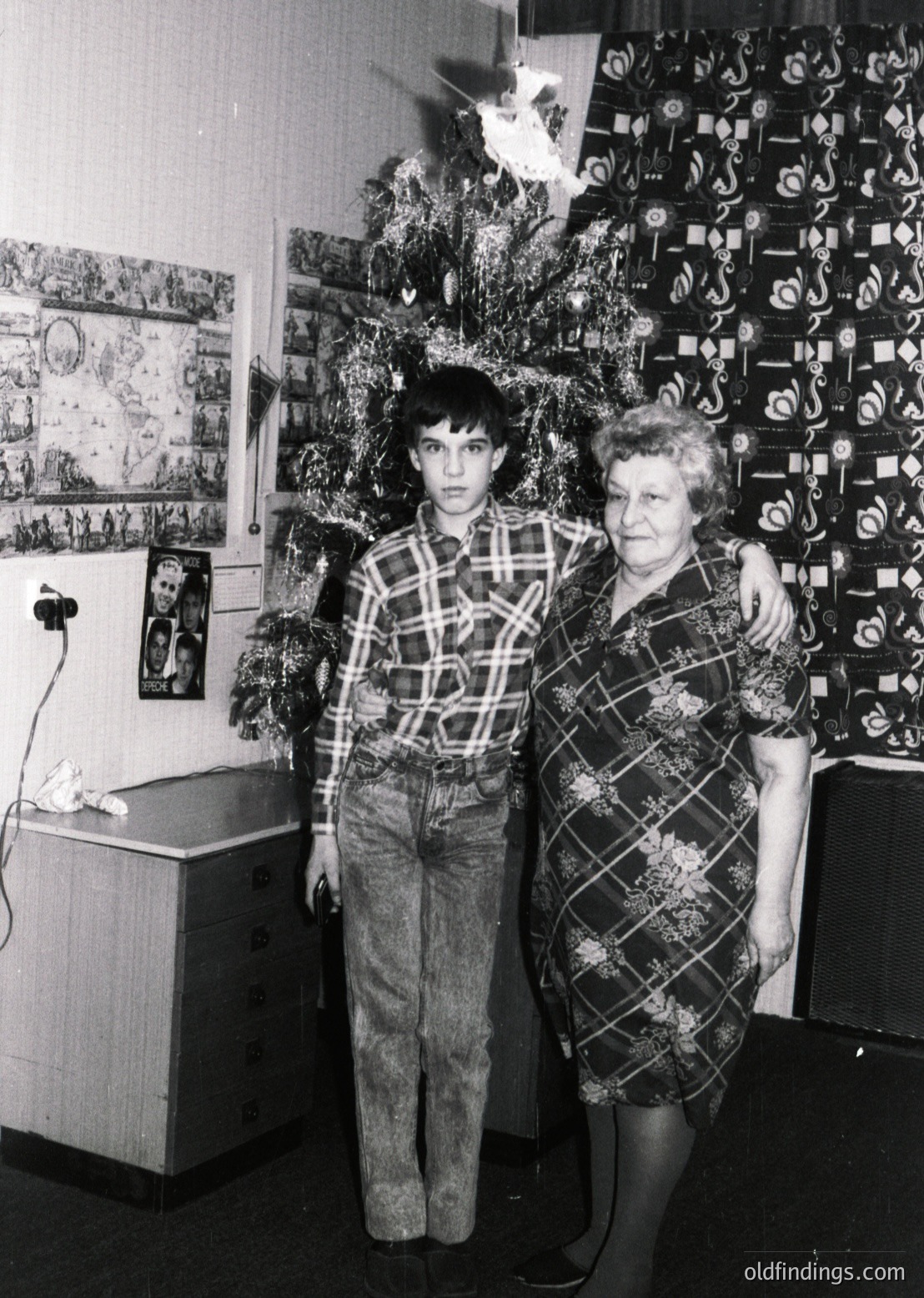 A black-and-white holiday portrait from the 1970s–80s, featuring a man and woman posing beside a decorated Christmas tree adorned with paper ornaments and a star topper. The woman wears a geometric-pattern dress, while the man sports a plaid shirt and bell-bottom jeans. A wall display of framed awards or certificates and a vintage radio or speaker in the background suggests a domestic setting with a focus on family and achievement. Ideal for vintage nostalgia, historical research, or stock imagery on family traditions.
