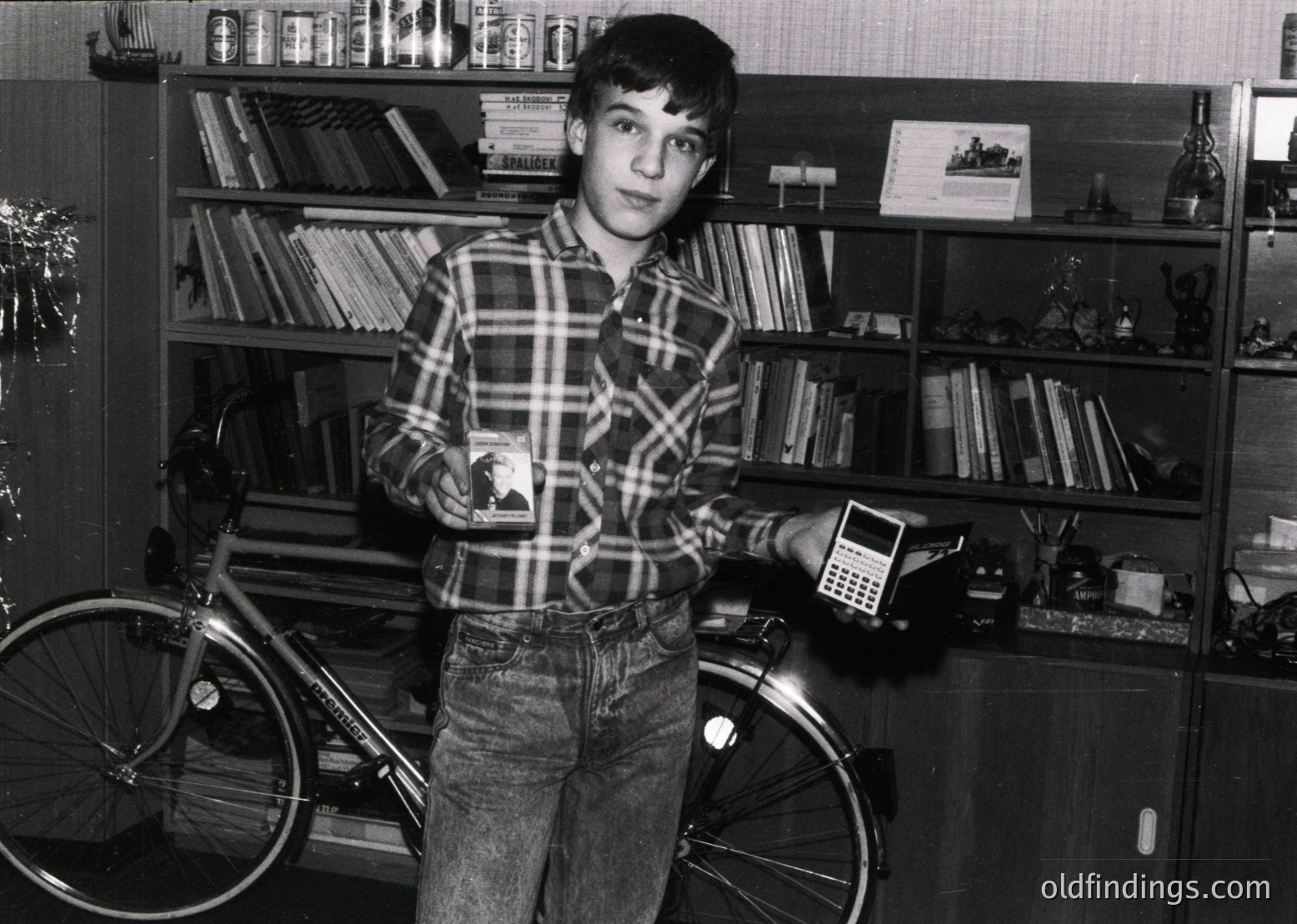 Mid-20th century boy poses indoors beside a vintage bicycle and early calculator. Bookshelves filled with hardcover volumes and decorative items frame the scene. Plaid shirt and jeans suggest 1970s–1980s Western household. Ideal for historical research or nostalgic design references.