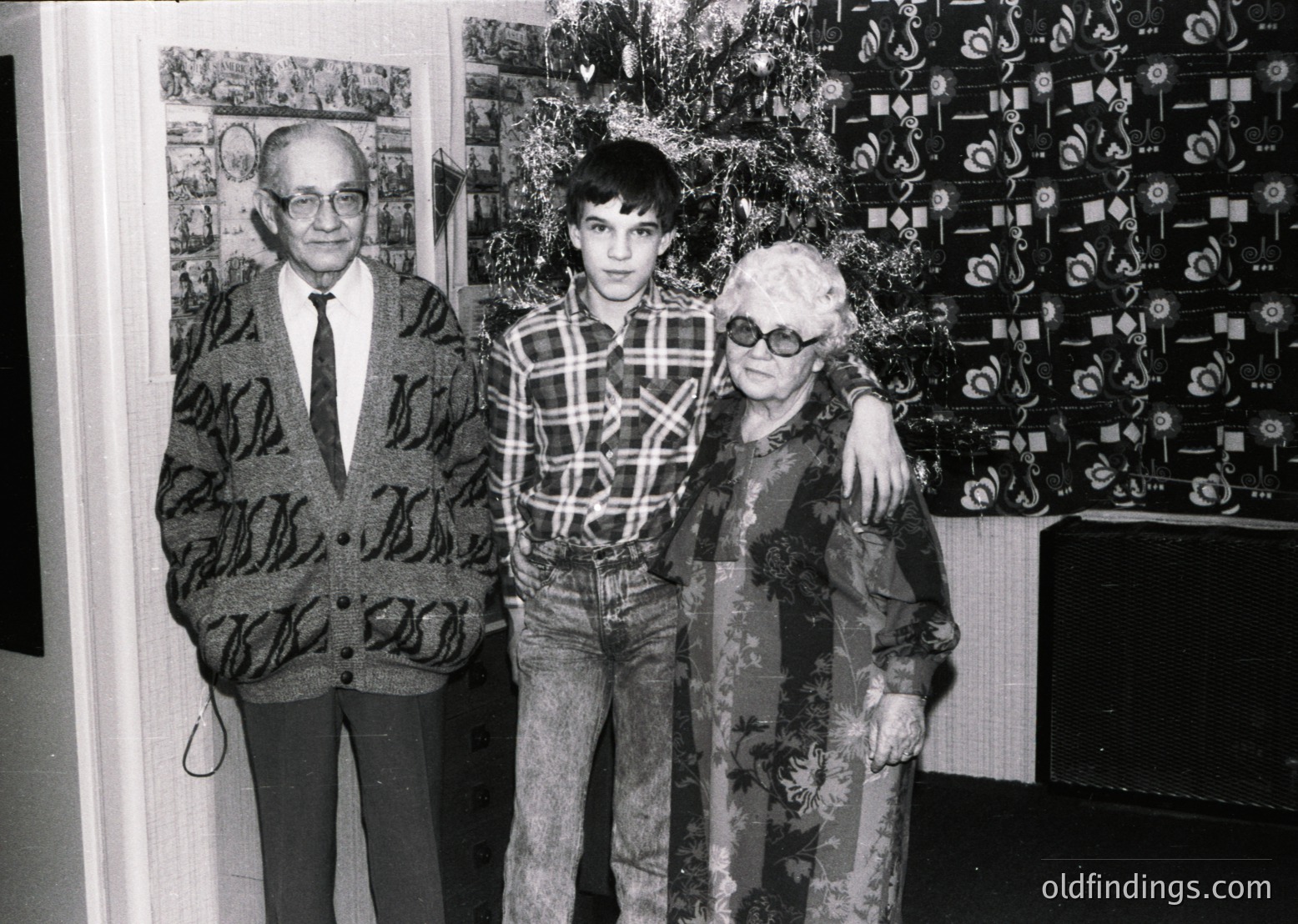 Intergenerational portrait featuring a man in a patterned blazer and glasses, a young boy in a plaid shirt, and an elderly woman in a floral-patterned coat. Backdrop includes framed artworks and a decorated Christmas tree with geometric ornaments. Likely 1970s–1980s, Western Europe/USA.